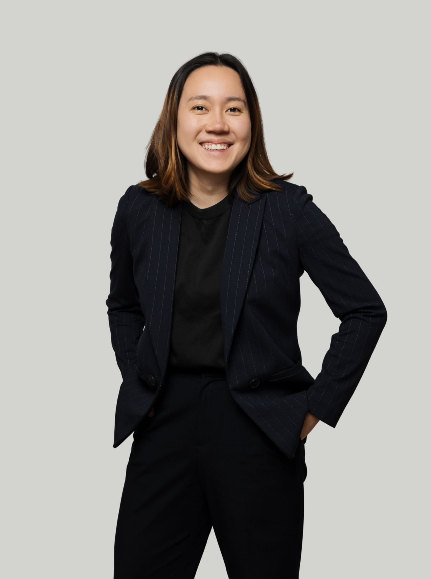 A woman with shoulder-length brown hair smiling, wearing a dark pinstripe suit and black shirt, standing against a plain light-colored background.