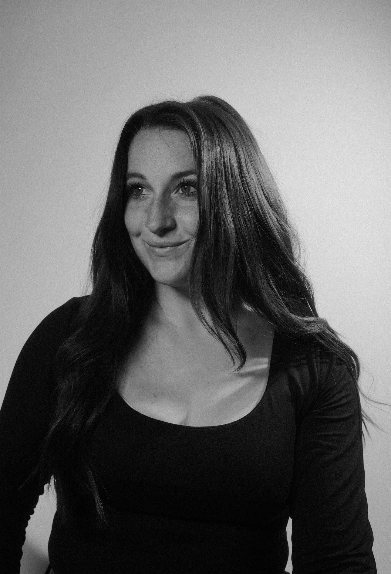 A black and white portrait of a woman with long, wavy dark hair, wearing a scoop-neck top, smiling softly and looking slightly to her right against a plain light-colored background.