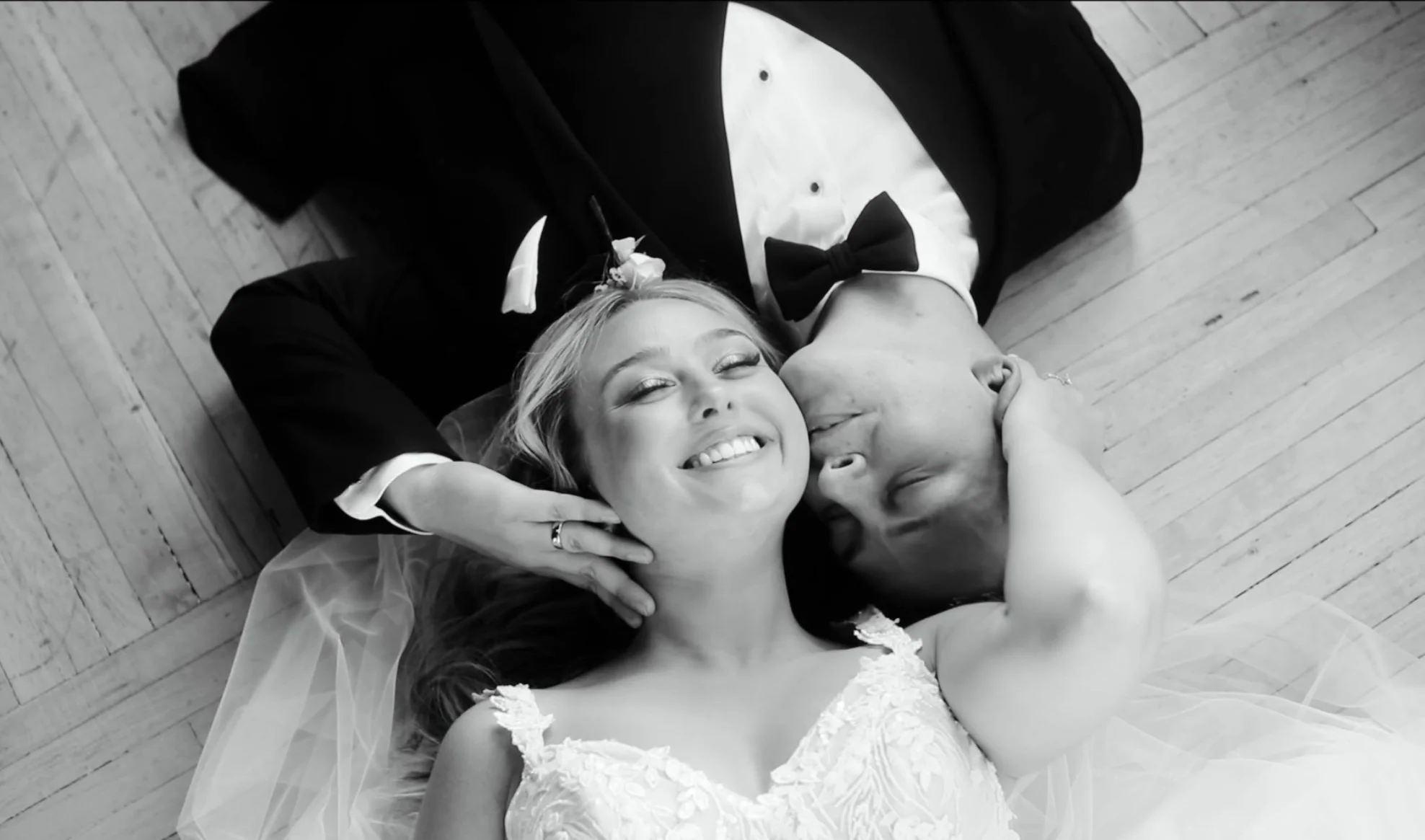 A man and woman in wedding attire lying on a wooden floor, smiling with eyes closed, with the woman in a white wedding dress and the man in a black tuxedo, sharing an intimate moment.