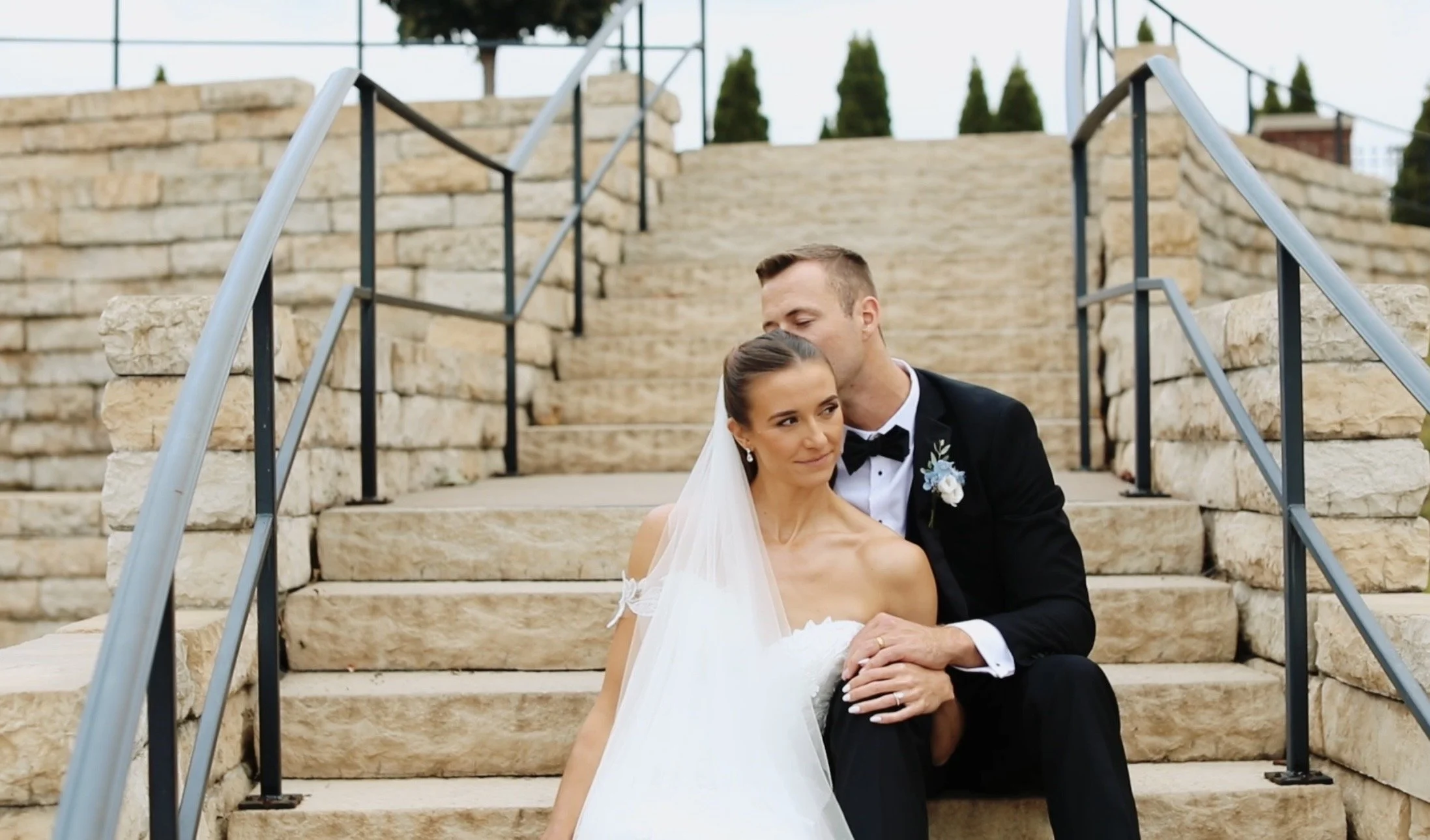 A bride and groom sitting on stone steps outdoors, with the groom kissing the bride's forehead. The bride is wearing a white wedding gown and veil, and the groom is in a black tuxedo with a boutonniere. Stone steps and a metal railing are visible in the background.