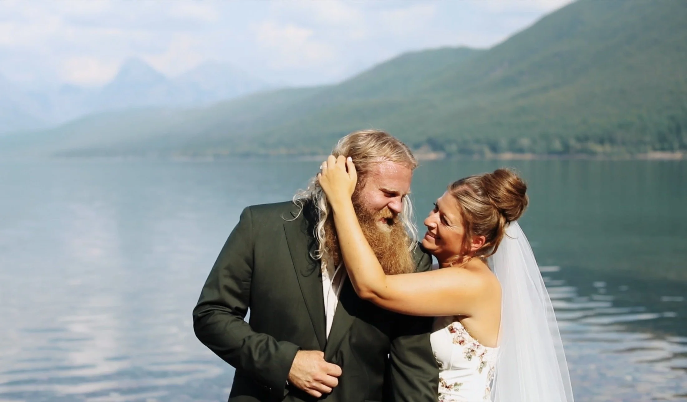 A couple dressed in wedding attire sharing a joyful moment by a lake, with mountains in the background.