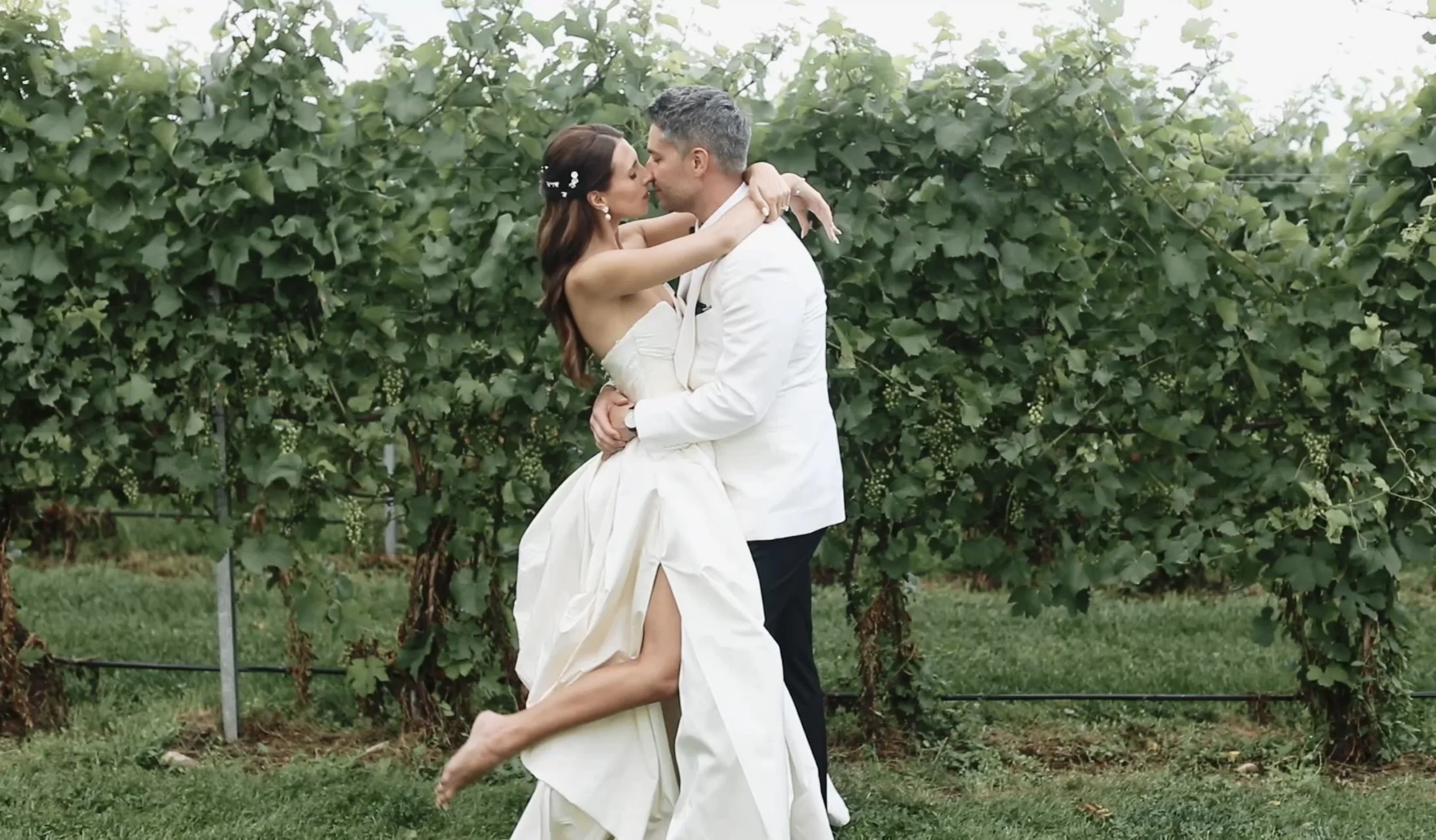 A bride and groom in wedding attire embracing and about to kiss in a vineyard with green leaves in the background.