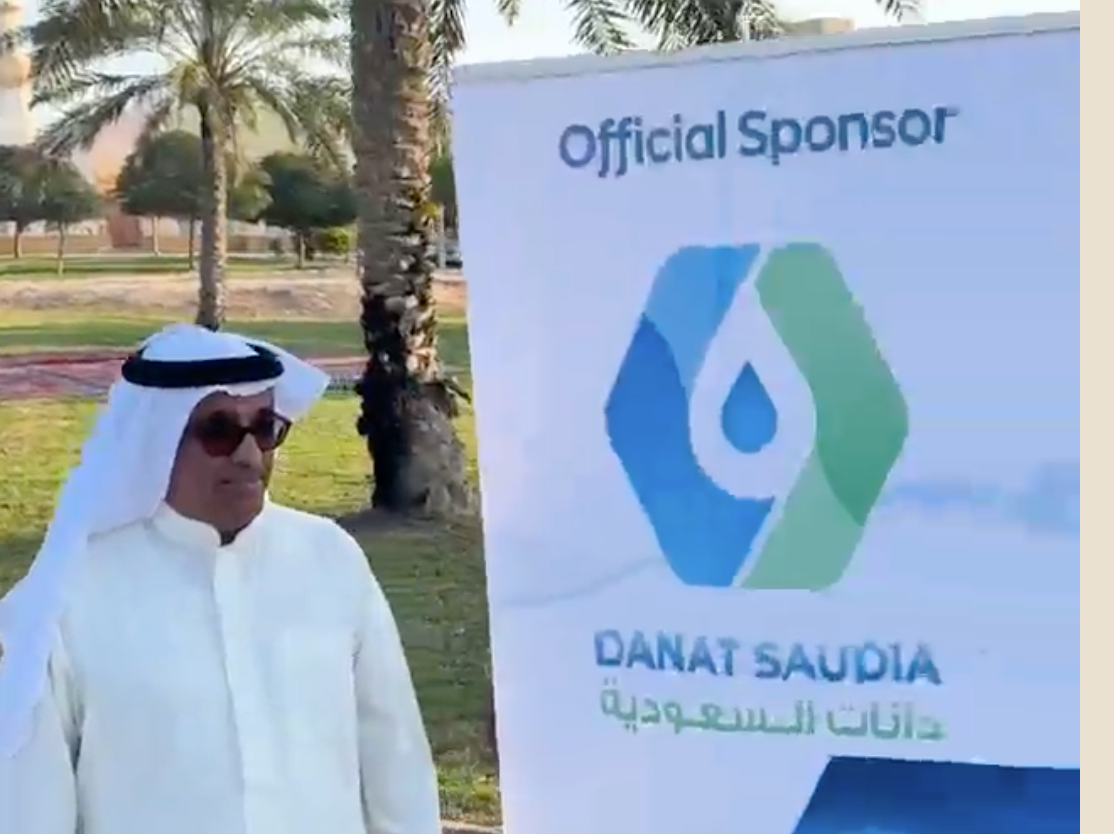 A man in traditional Middle Eastern attire and sunglasses standing outdoors next to a sign that reads 'Official Sponsor' with a logo and the text 'DANAT SAUDIA' in English and Arabic, with palm trees and a park in the background.