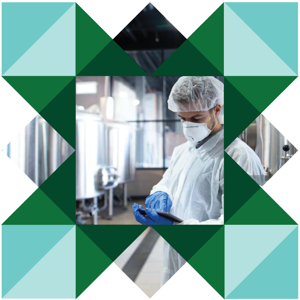 A person wearing a lab coat, gloves, a face mask, and a hair net in a brewery or distillery, looking at a mobile device.