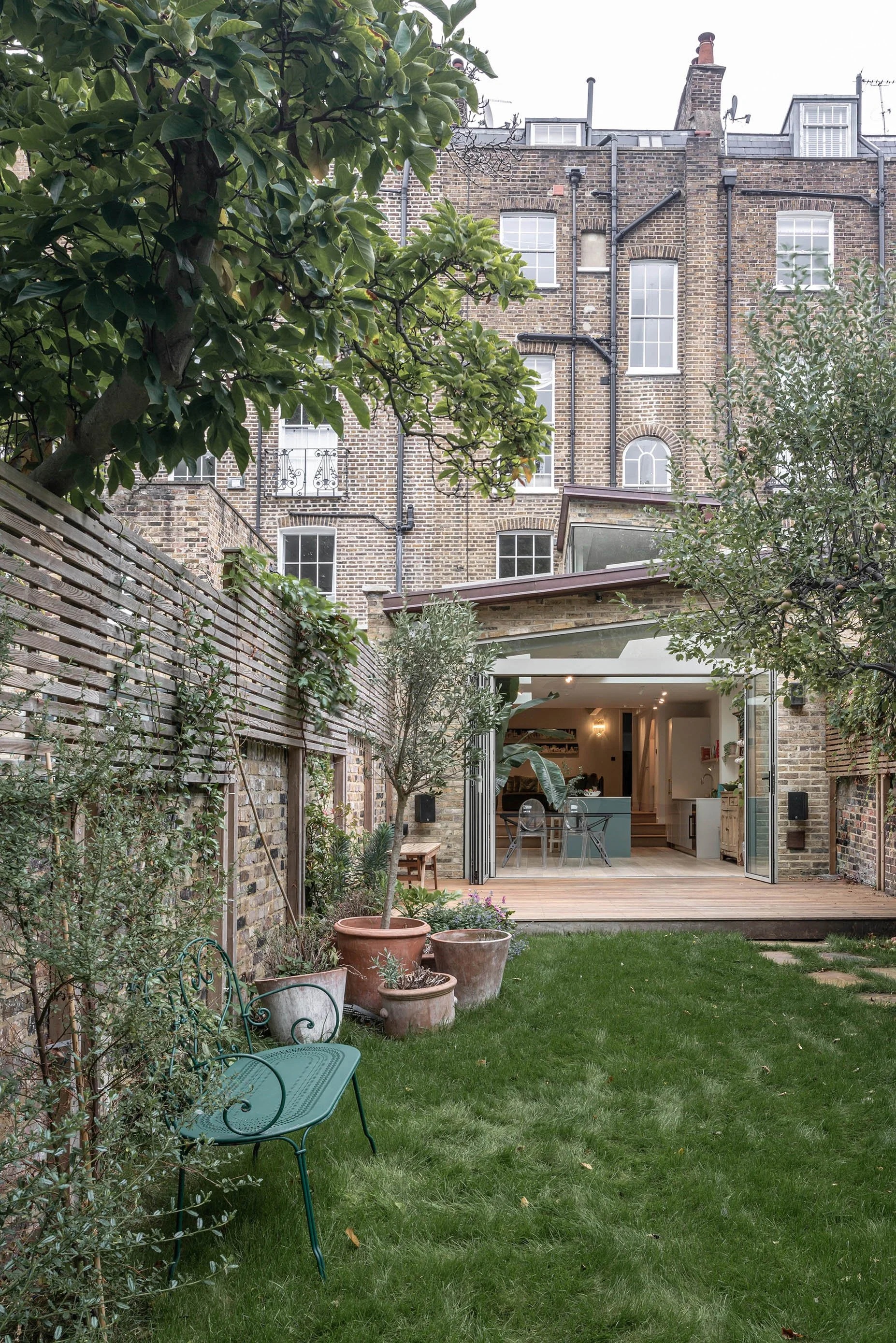 Rear view of a London terrace house with a copper-clad pitched roof extension, large sliding doors opening onto a timber deck, and a kitchen dining space connected to a planted garden.
