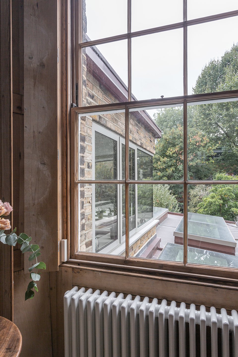 View through a timber sash window towards a brick rear extension with rooflights, framed by timber panelling and a radiator below.