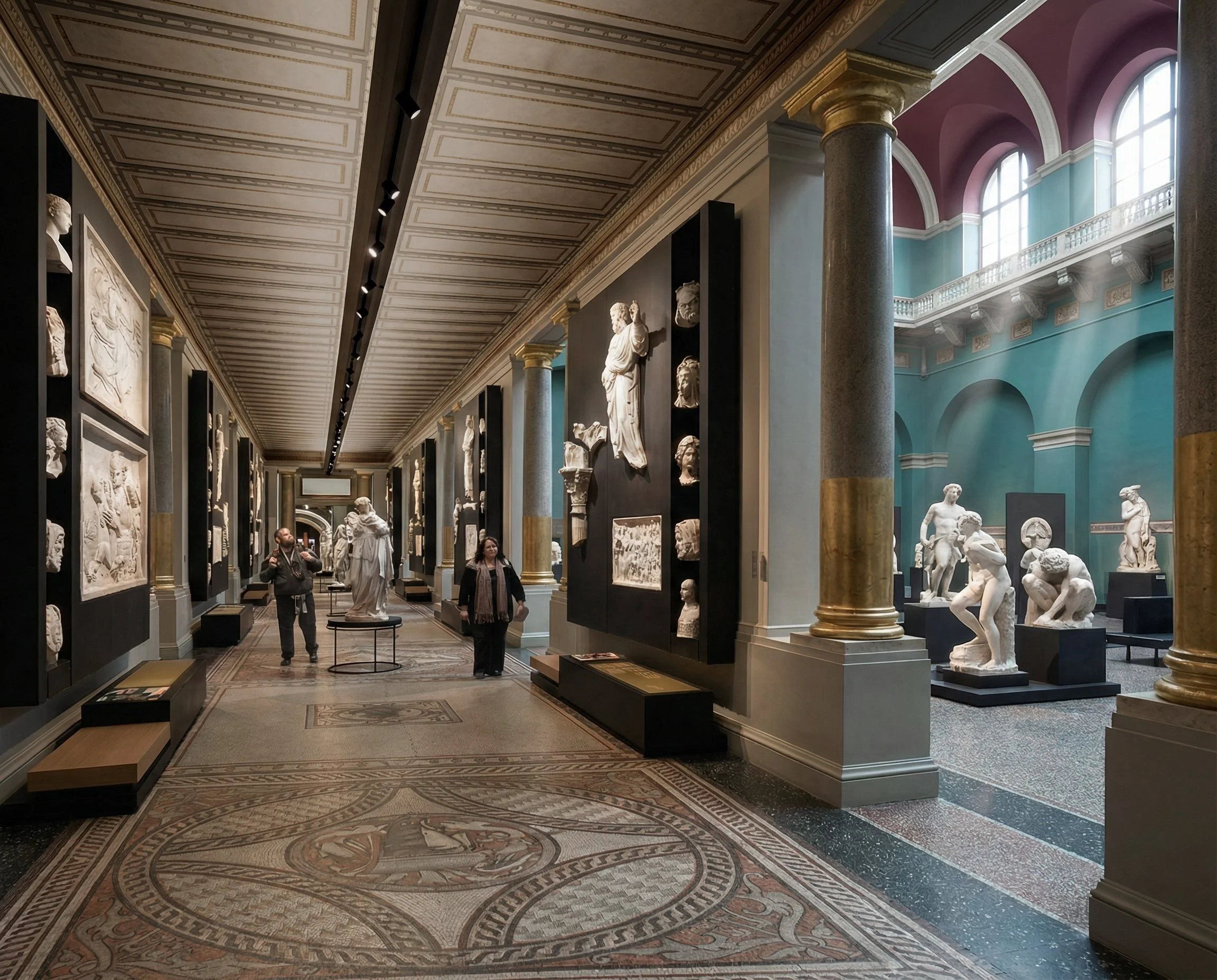Interior view of the Cast Courts showing a linear gallery with plaster casts displayed on dark panels between historic columns, visitors moving through the space, and adjoining galleries visible beyond.