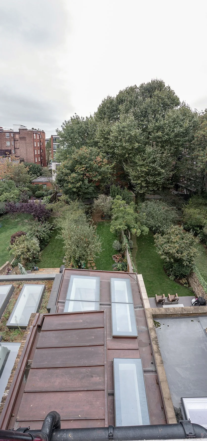 View over a copper roof with linear rooflights towards a landscaped garden and mature trees behind a London terrace.