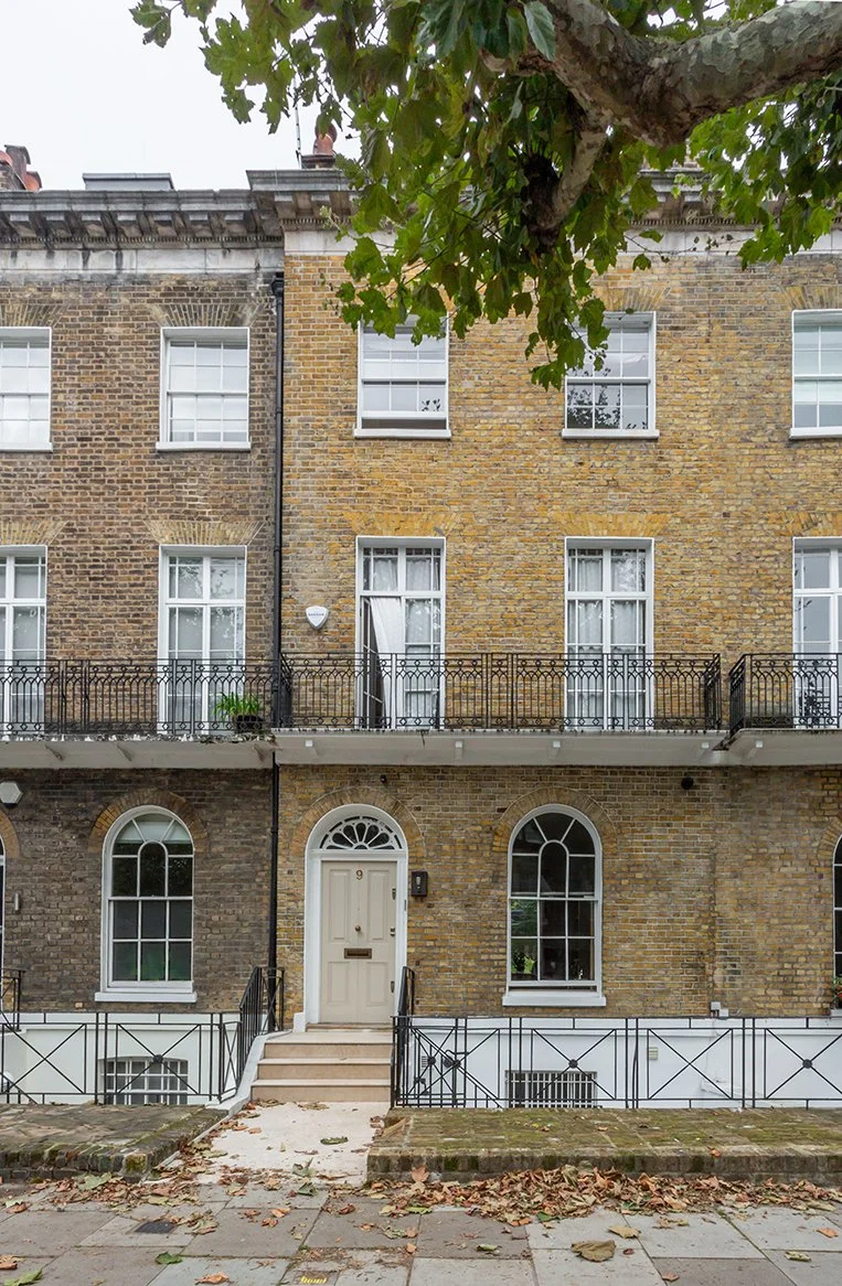 Front façade of a Grade II listed London terrace house in yellow stock brick with white sash windows, iron balcony, and central entrance under a fanlight.