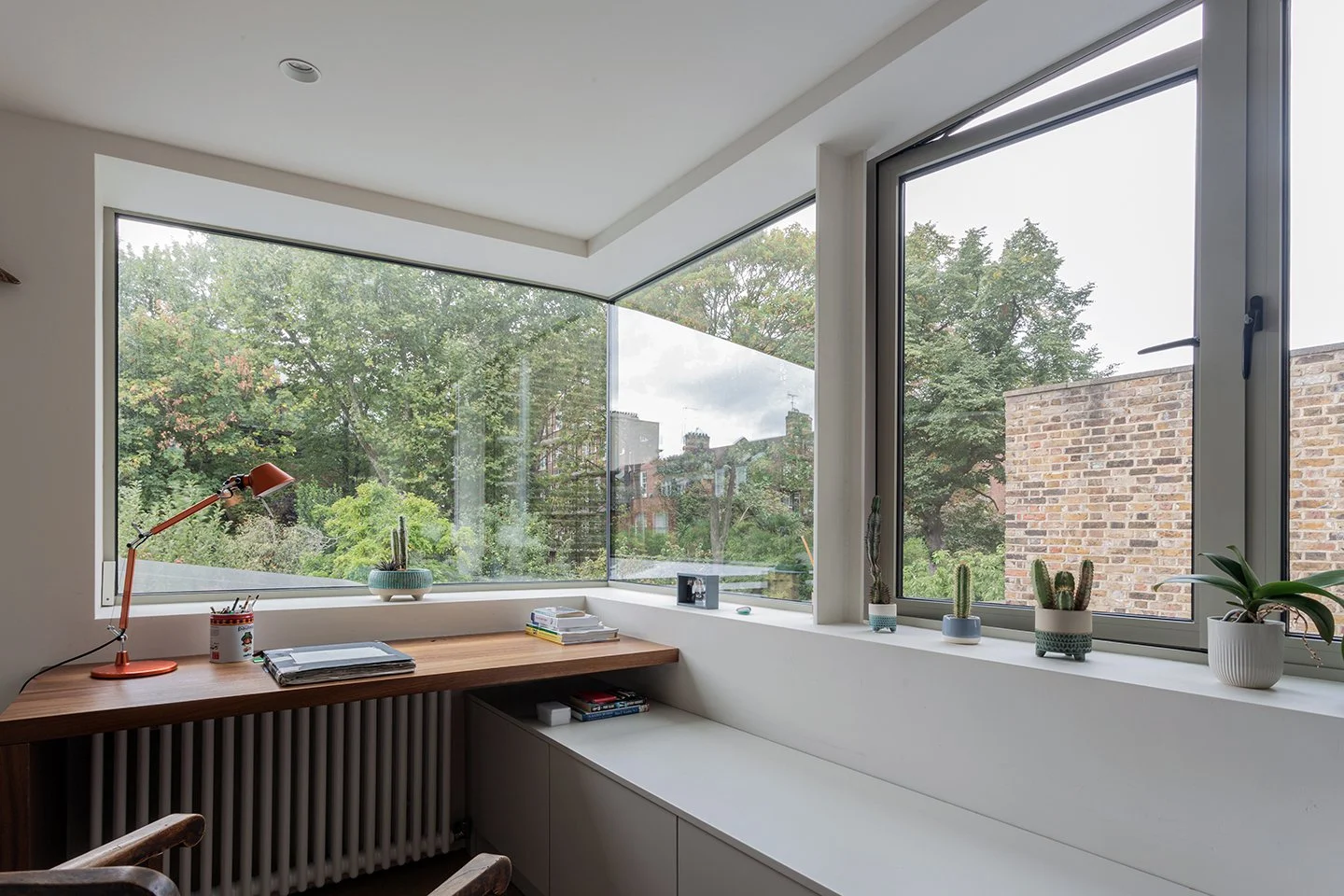 Corner study with wraparound glazing overlooking a garden, featuring a timber desk, built-in seating, and deep window reveals.