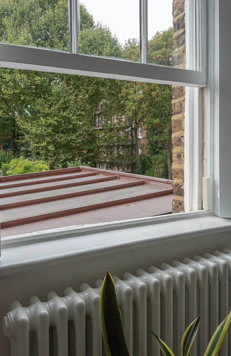 View from a sash window across a copper roof to a garden with mature trees, framed by painted timber joinery and a radiator below.