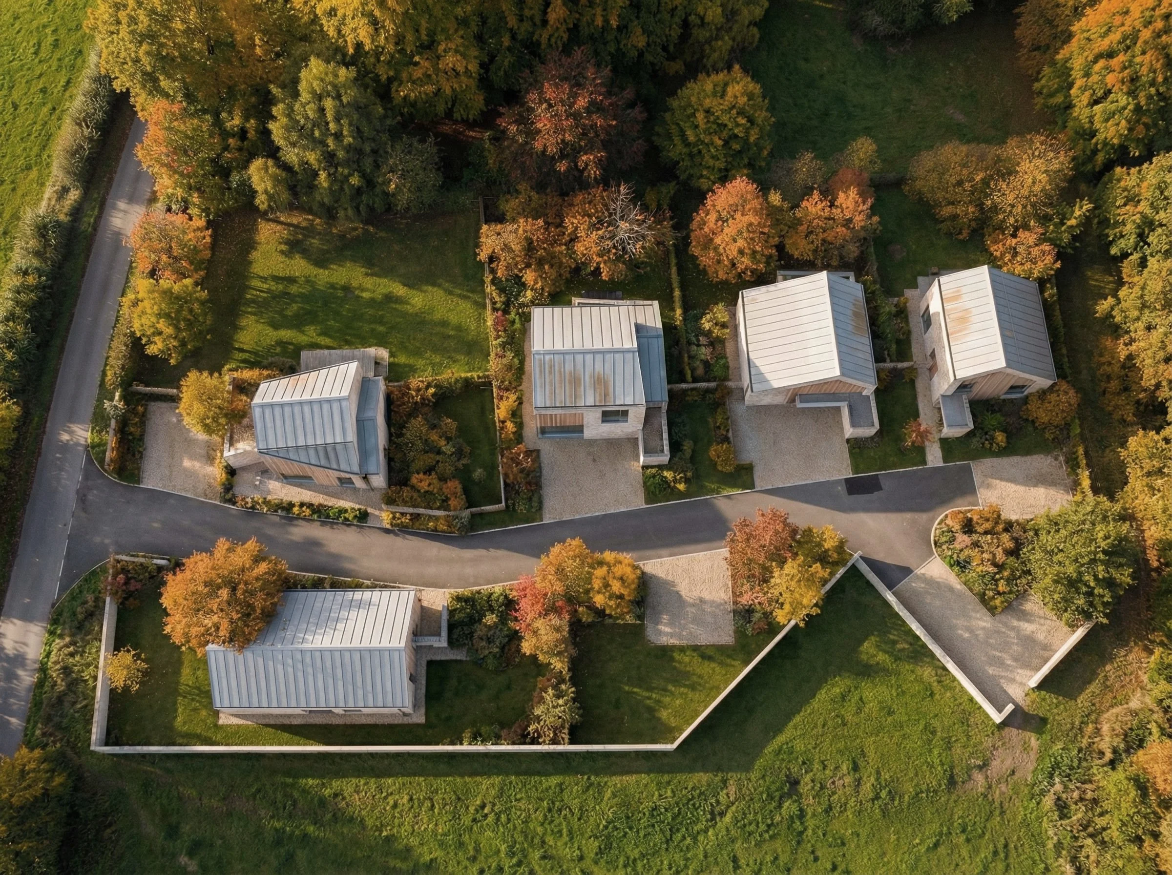 Aerial view showing six contemporary pitched-roof houses arranged along a gently curving internal road, set within landscaped gardens and mature trees, illustrating a landscape-led residential development.