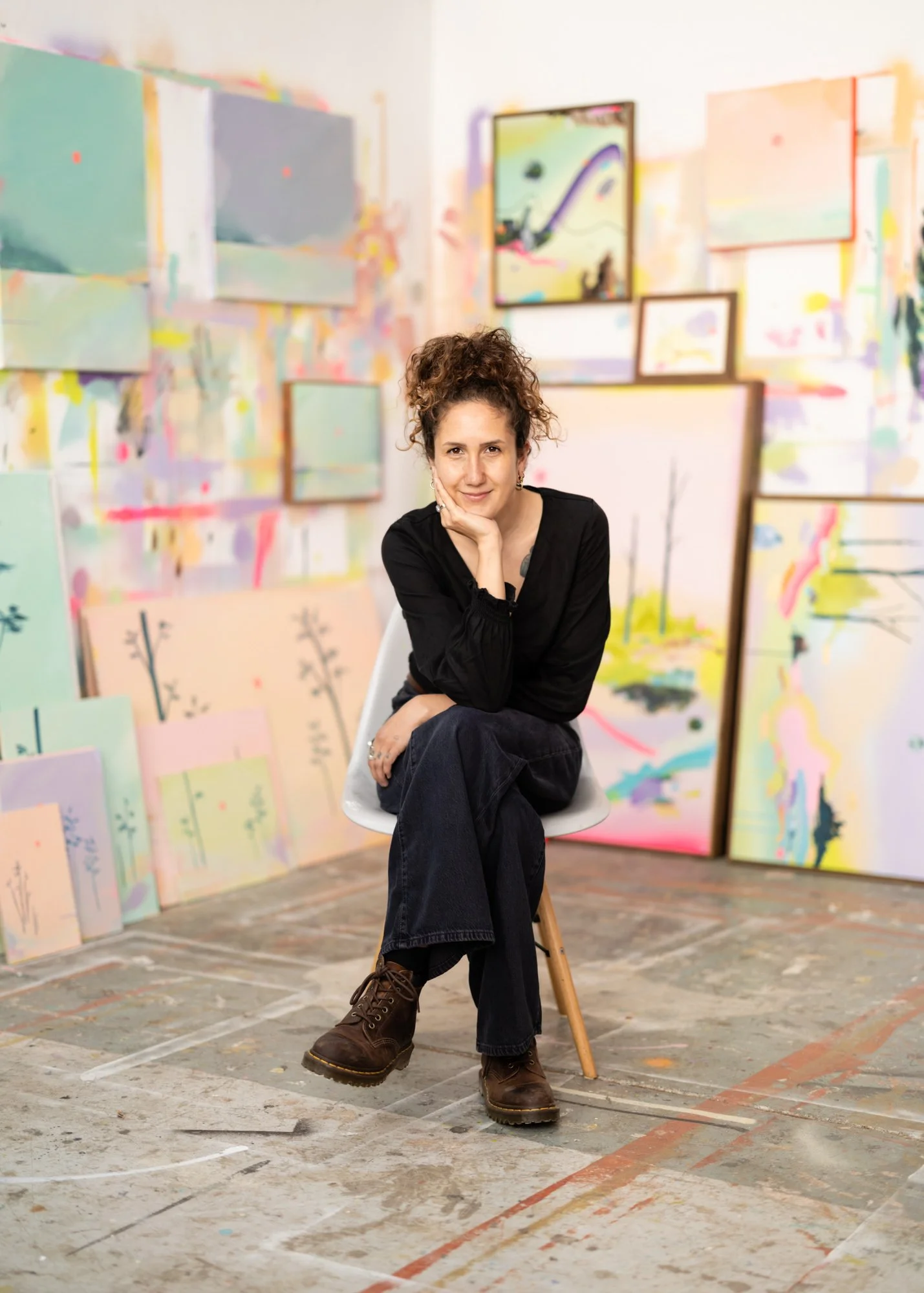 A young woman with curly hair sitting on a white chair in an art studio, surrounded by colorful abstract paintings and canvases leaning against the walls.