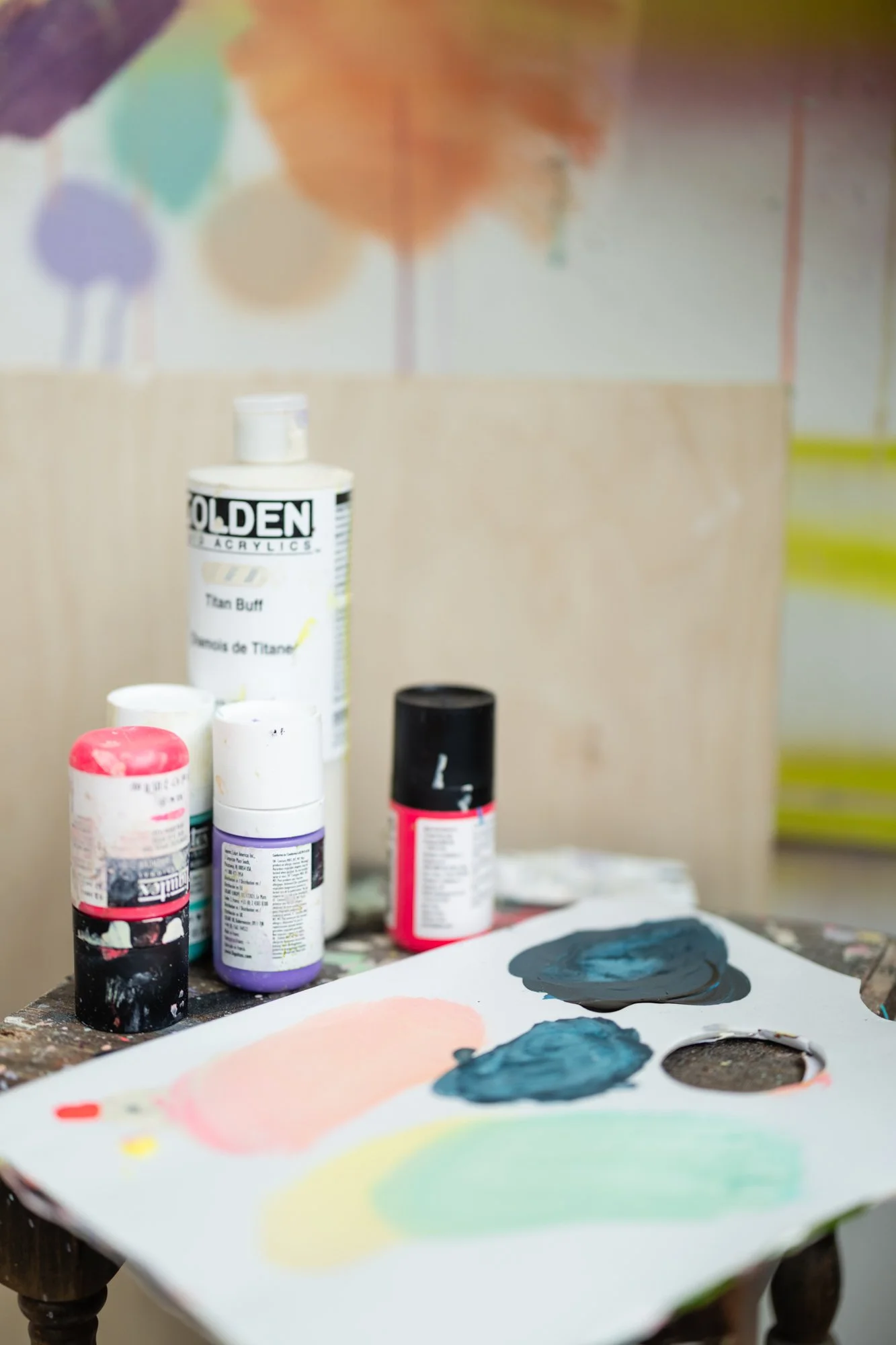 Art supplies on a wooden table, including bottles of acrylic paint, a paint palette with mixed colors, and a colorful abstract painting in the background.