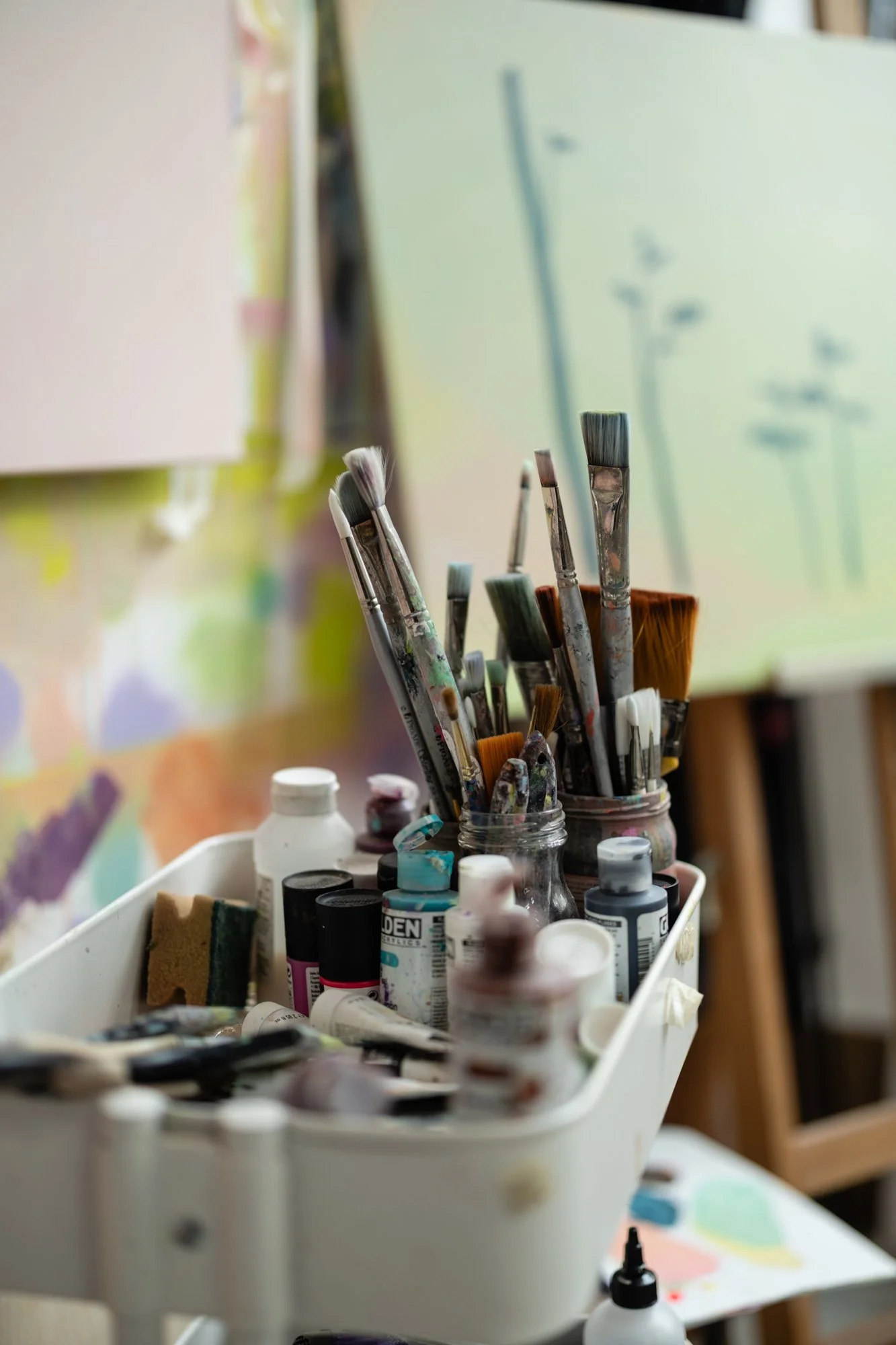 A collection of paintbrushes and acrylic paint bottles on a white tray, with an unfinished painting in the background.