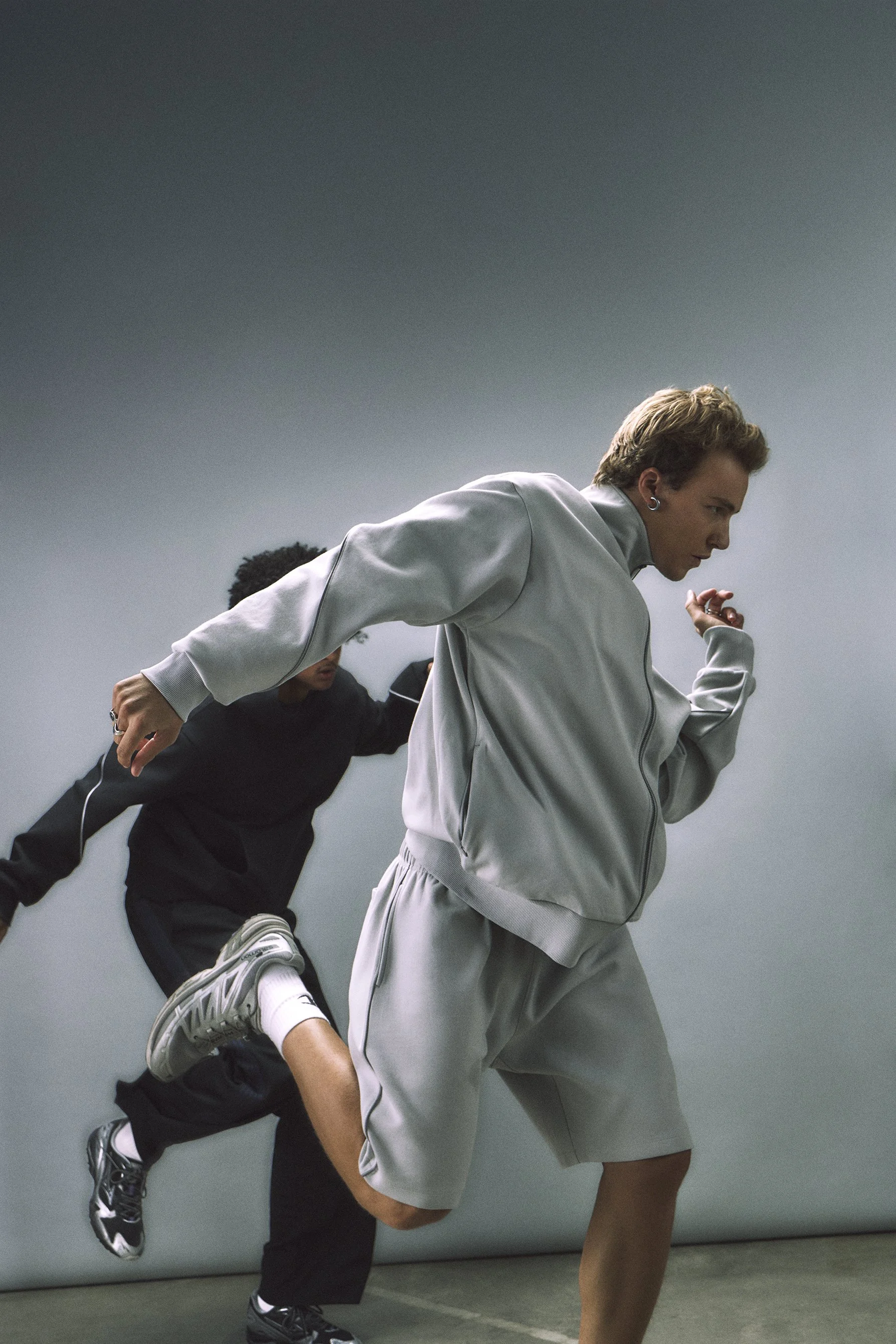 A young man with curly hair wearing a gray hoodie and matching sweatpants touching his hair, standing next to a young woman with wavy hair tied back, wearing a white cropped sweatshirt and black pants, both against a plain gray background.