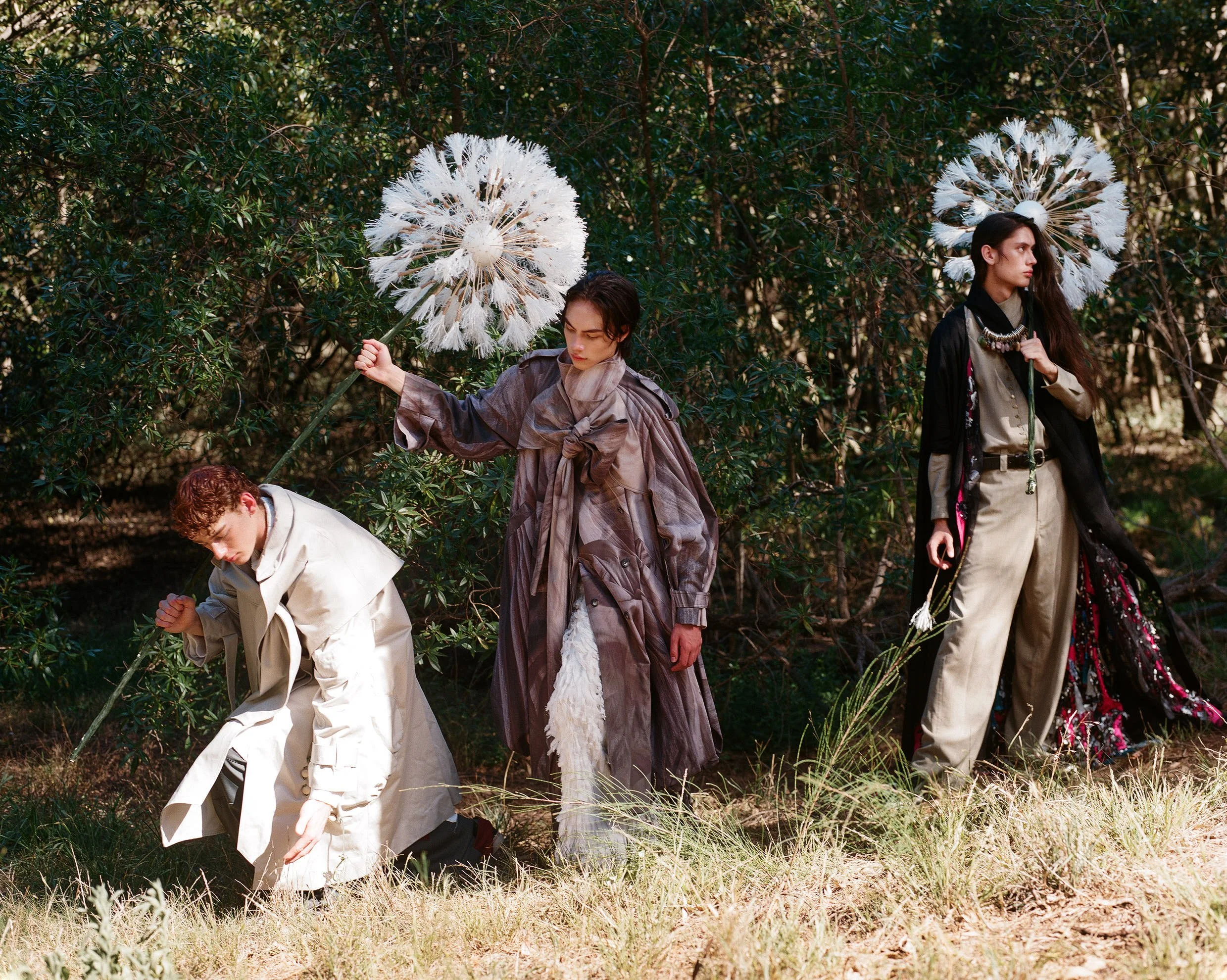 Three models standing outdoors in a forest, dressed in fashionable clothing, holding large white dandelion-like props, with trees and grass in the background.