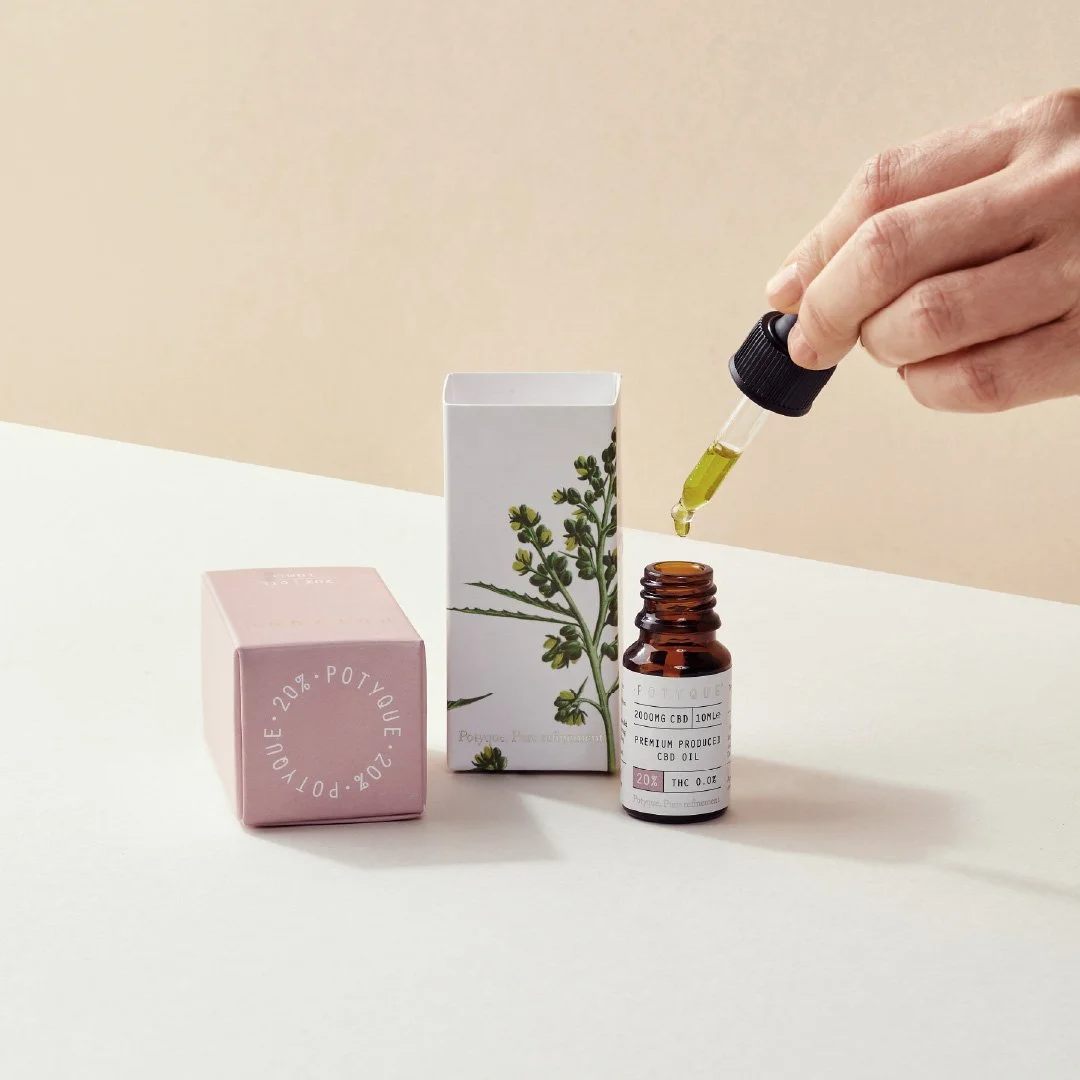 A hand holding a dropper releasing CBD oil into a small amber glass bottle, with a white box with plant illustration and a pink box on a white surface.