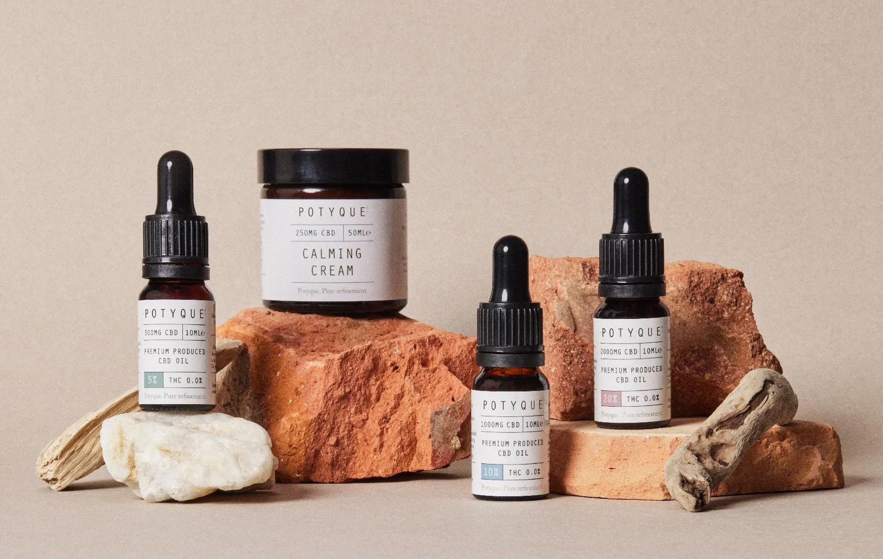 A collection of Potyque CBD oil bottles and a jar of calming cream arranged on bricks and pebbles against a neutral background.