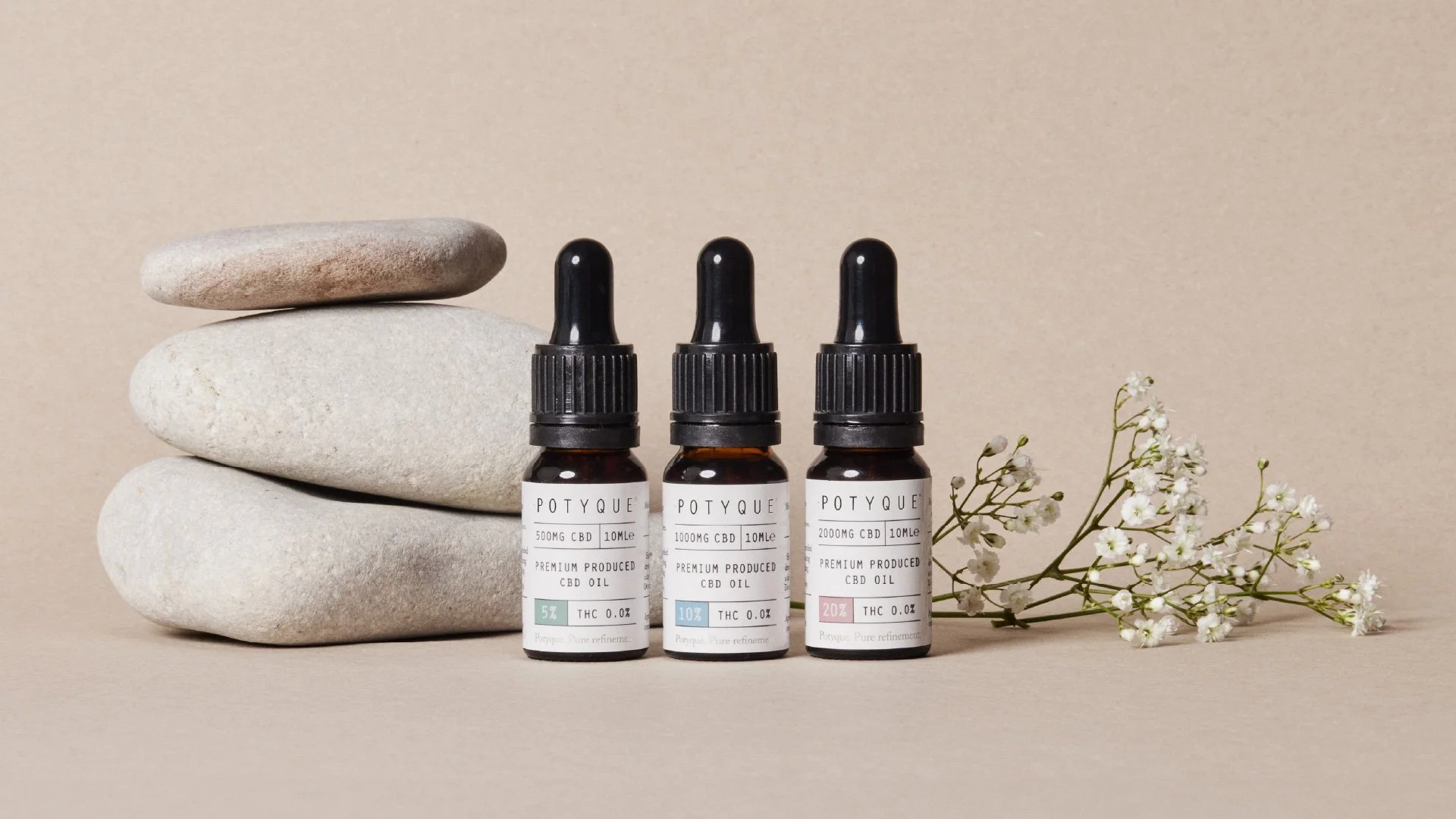 Three bottles of Potyque CBD oil labeled with different concentrations, stacked stones, and white flowers against a beige background.