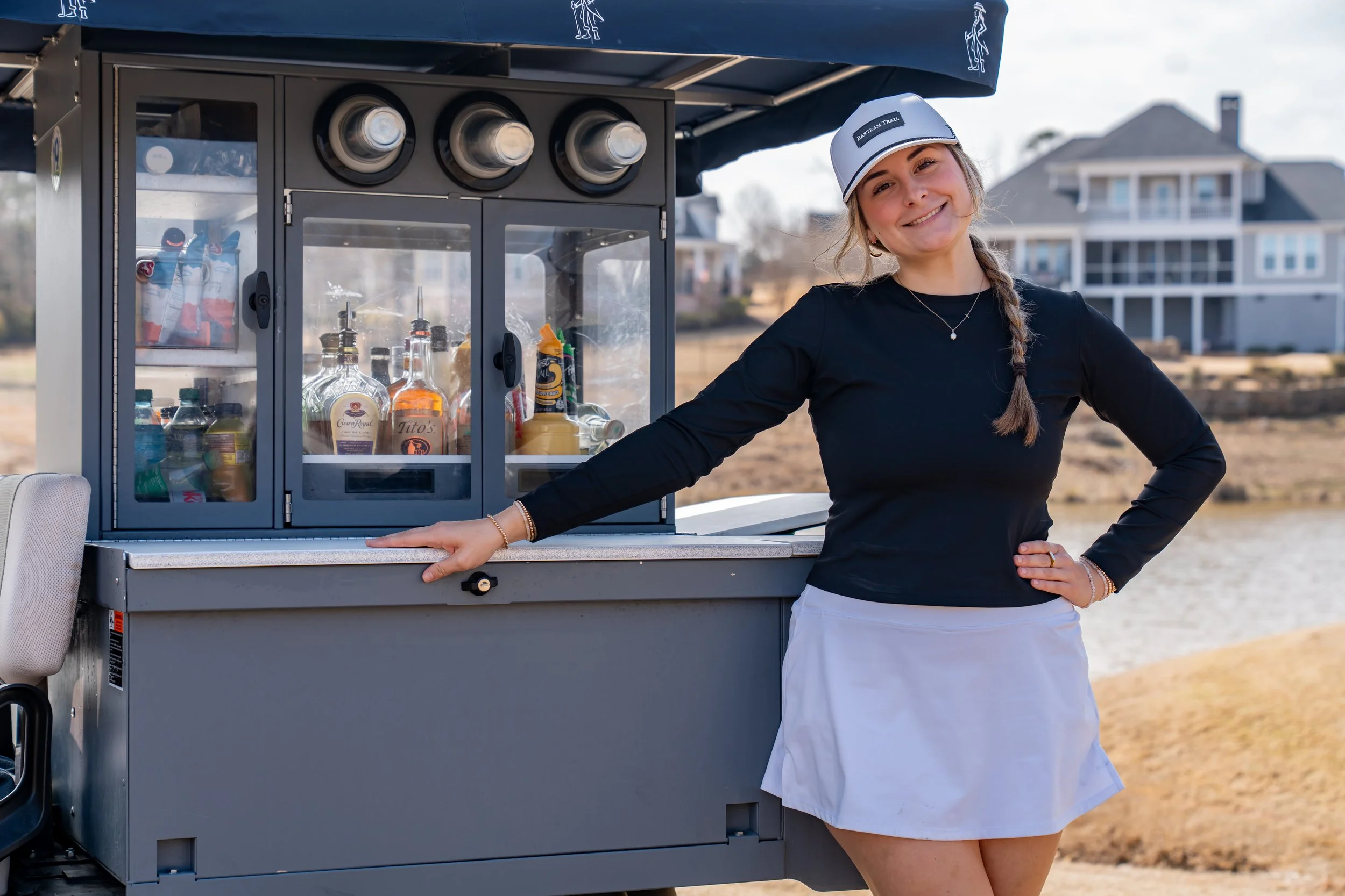 A young woman wearing a black long-sleeve shirt, a white skirt, and a cap stands next to a mobile drink cart with bottled alcohol and condiments, smiling outdoors near a body of water with houses in the background.
