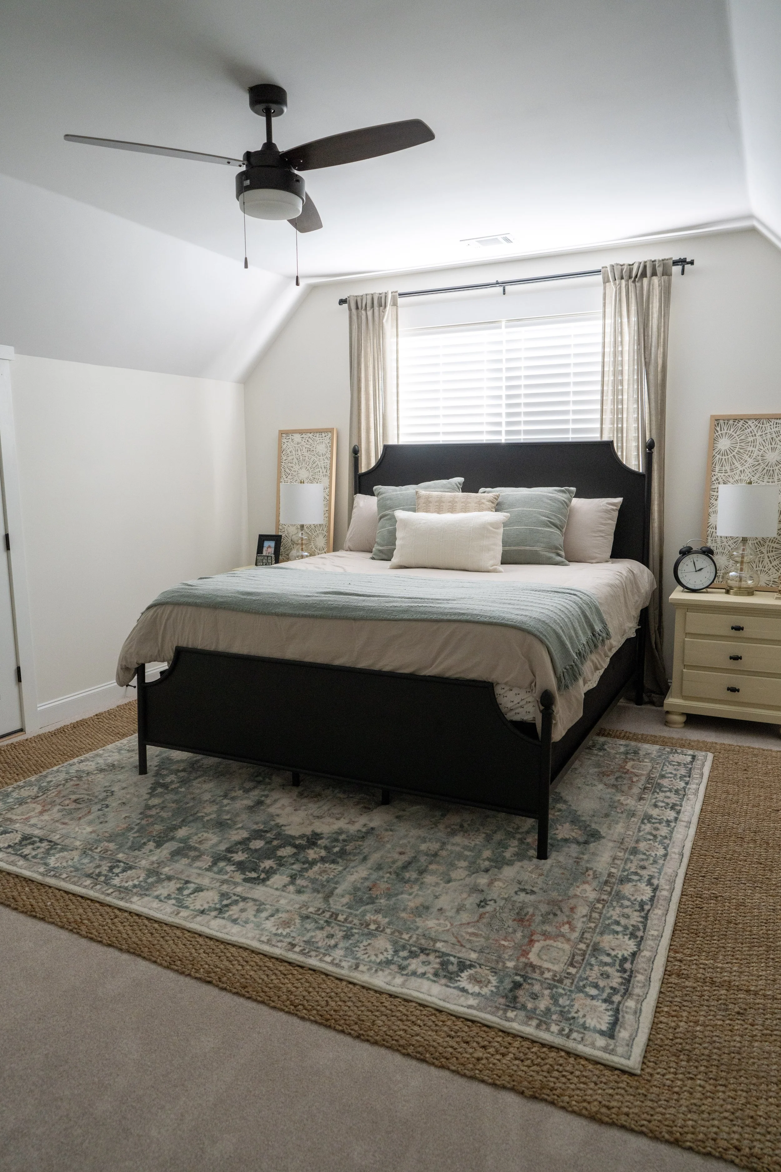 A bedroom with a black bed frame, light-colored bedding, and an area rug. There are beige drapes over a window, with a ceiling fan overhead. A nightstand with a clock and lamp is beside the bed.
