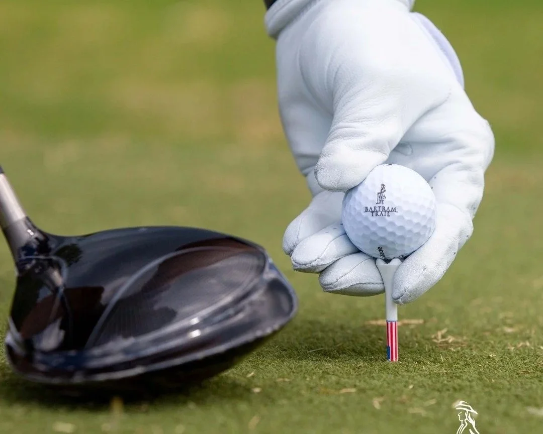 A person wearing a white golf glove placing a golf tee with a golf ball on it on the grass. The golf ball has the club's logo and the words "BARTRAM TRAIL" printed on it, with a golf club head nearby.