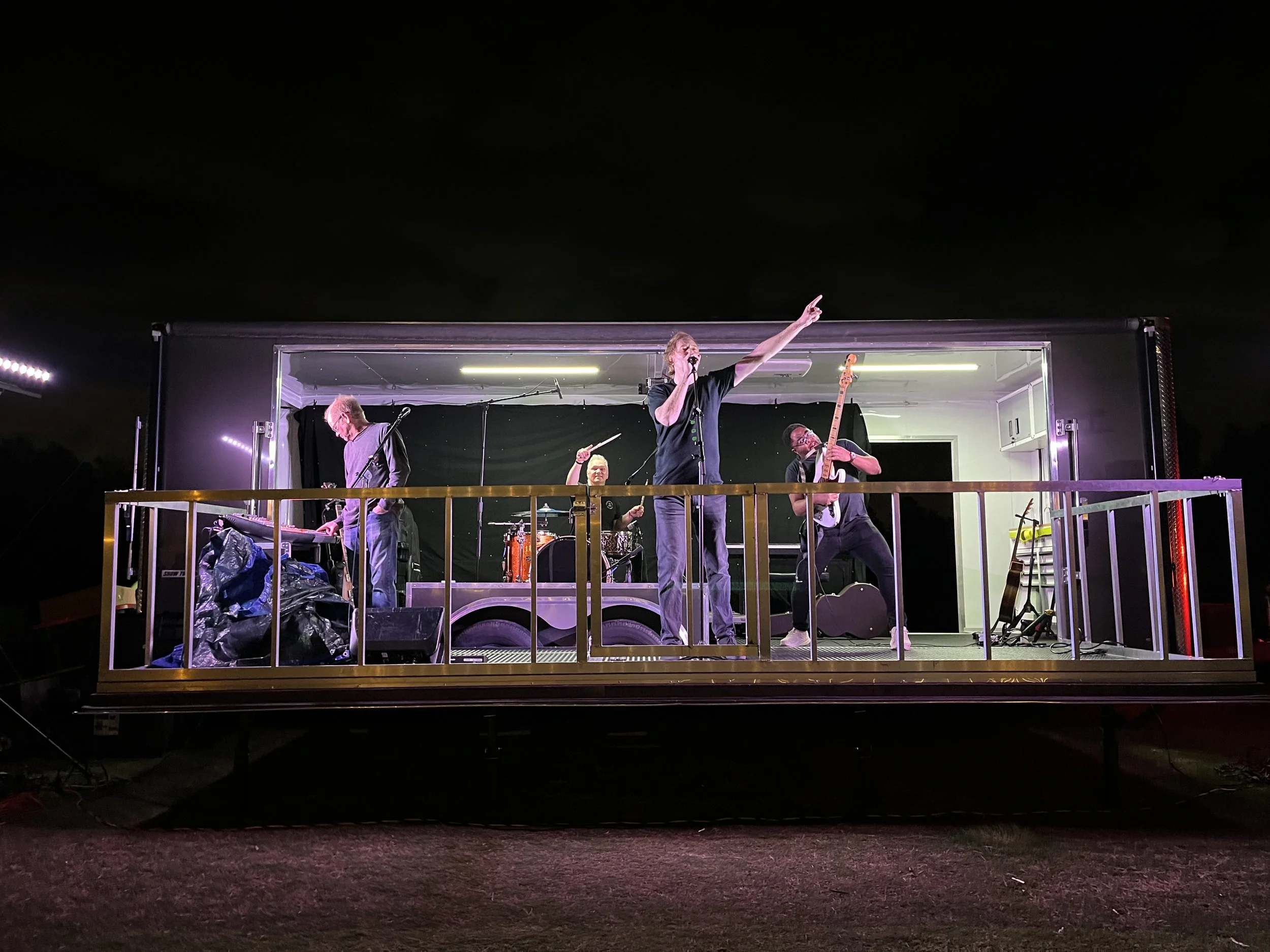 A band of five musicians performing on a stage inside an open trailer at night, with instruments including drums, guitar, and microphone, and stage lighting overhead.