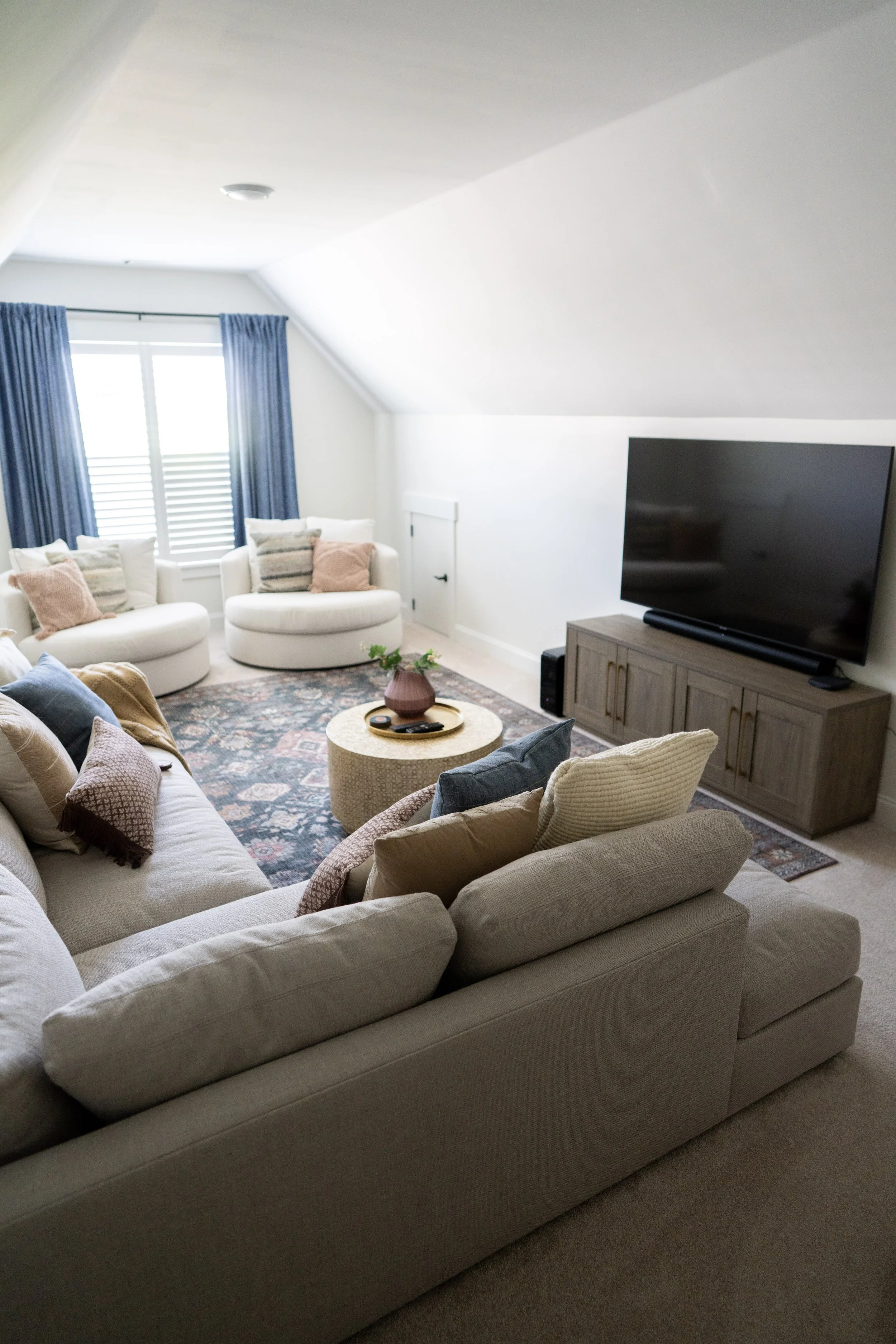 Cozy living room with white sectional sofa, blue curtains, patterned area rug, wall-mounted TV, and a coffee table with a pink vase and remote controls.