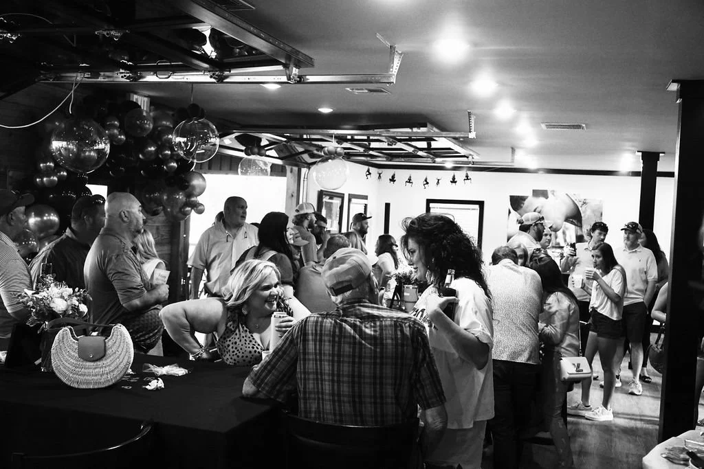 People mingling at a social gathering or party, with some holding drinks in a decorated indoor venue featuring balloons and festive decorations.
