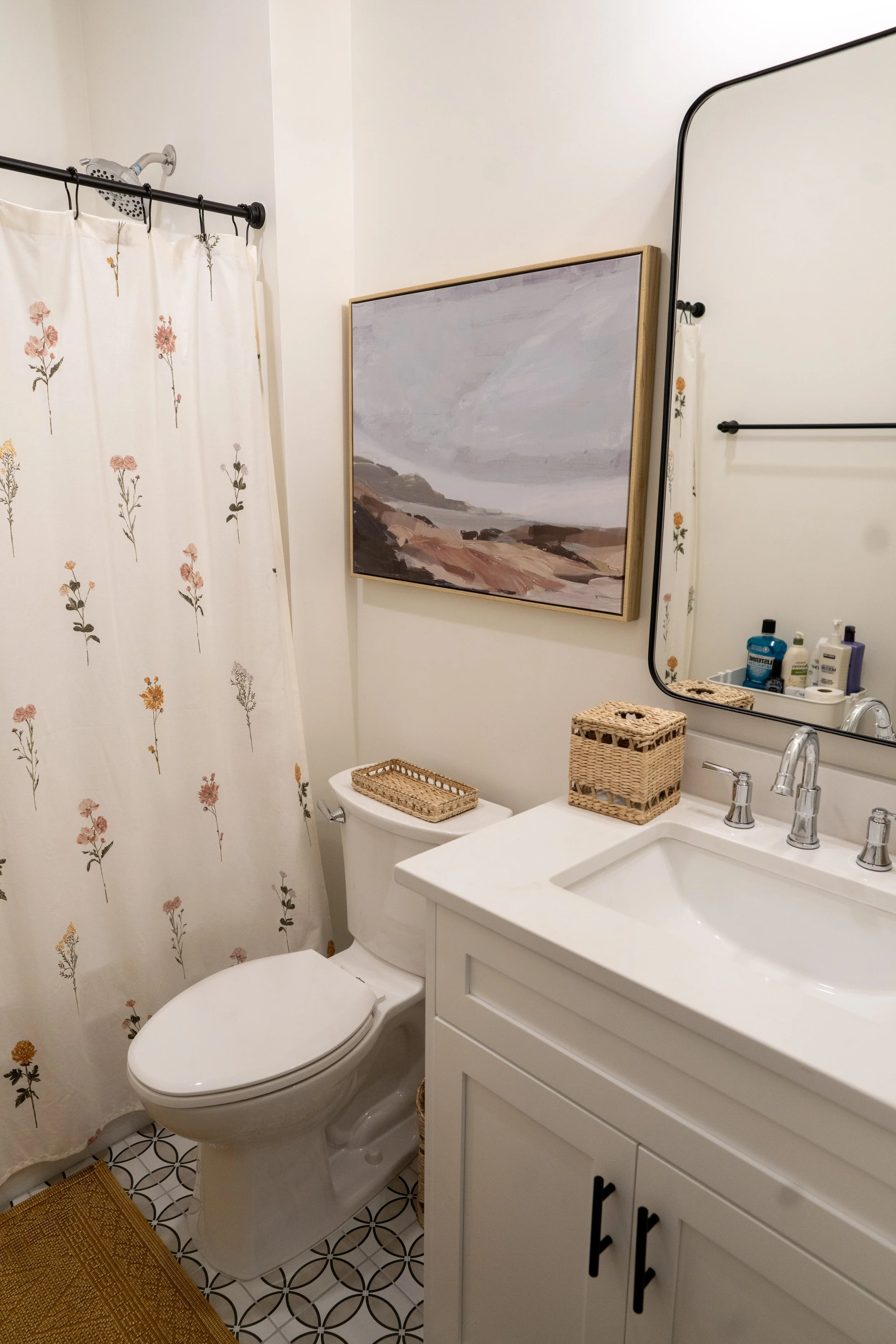 A bathroom with a shower curtain decorated with pink and yellow flowers, a white toilet with a wicker tray on top, a white vanity with a sink, a large mirror, a framed landscape painting, and several toiletry bottles on the counter.