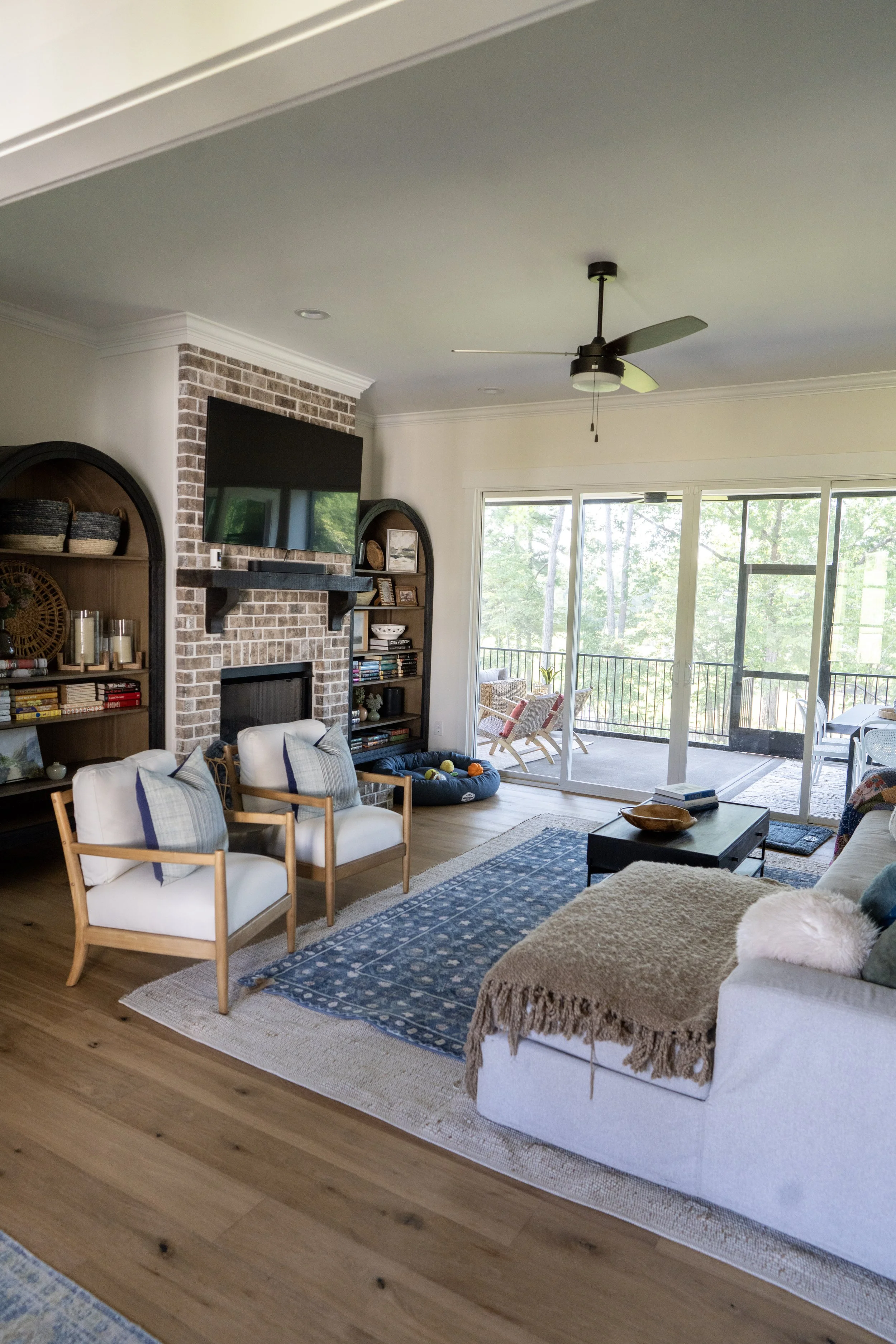 Living room with chairs, a sofa, a coffee table, bookshelves, a brick fireplace with a mounted TV, and large glass sliding doors leading to a balcony with outdoor seating.