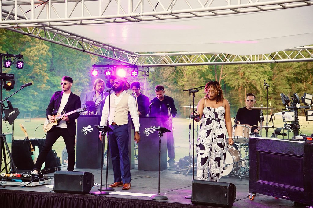 A band performs on an outdoor stage with five members: a guitarist, a vocalist, a keyboardist, a drummer, and a bassist, with trees in the background.