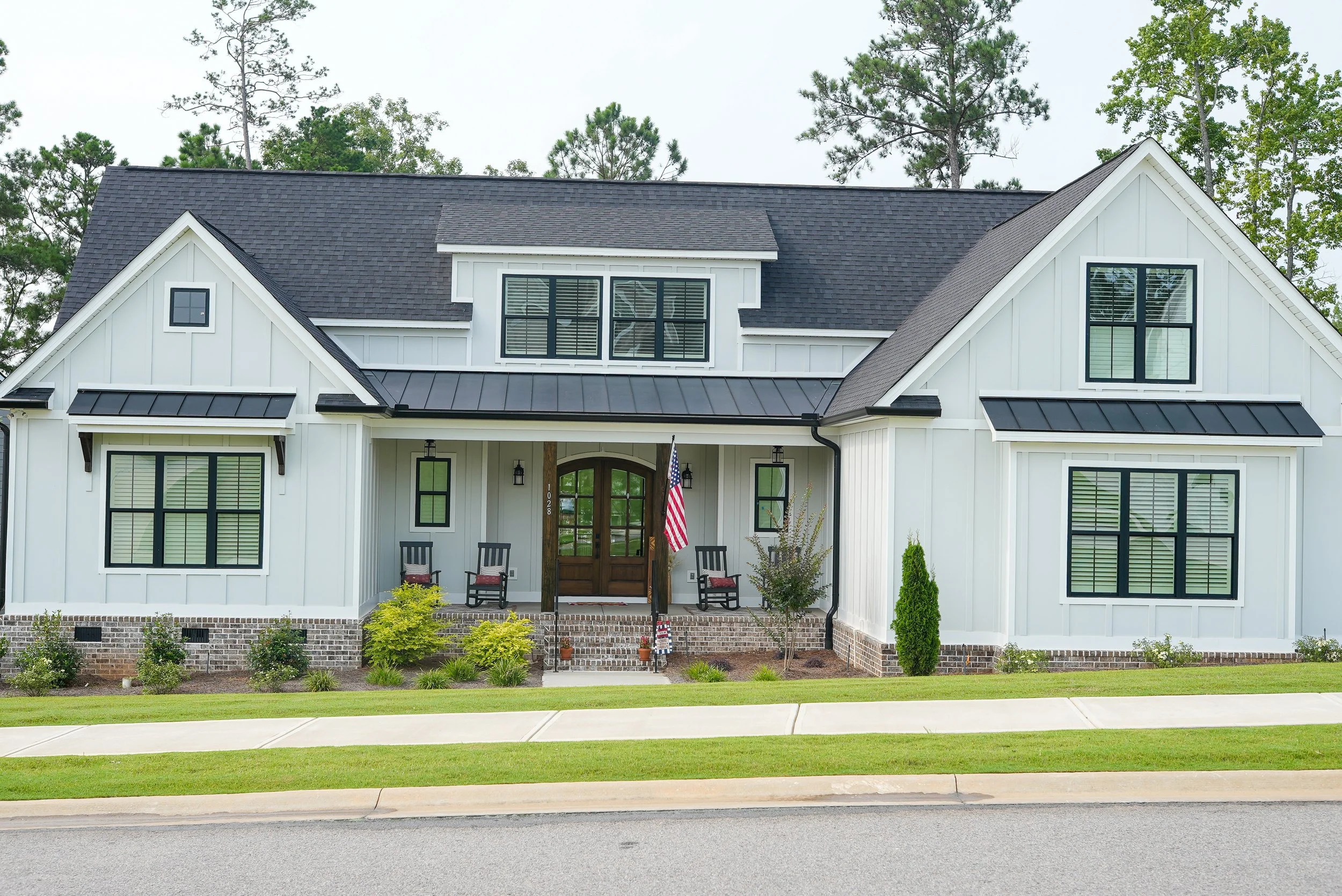 A modern two-story white house with black window frames, a gabled roof, and a front porch with two black rocking chairs, an American flag, and landscaping with small bushes and grass.
