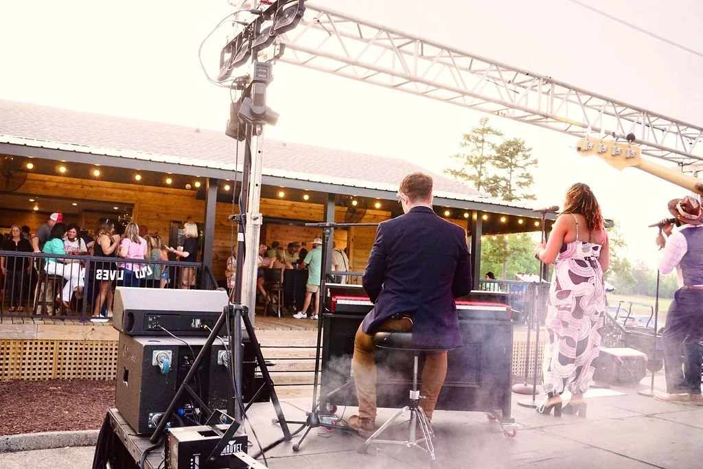 A live outdoor music performance at a social event, with musicians playing on stage and people gathering nearby, some wearing festive attire like Santa hats.