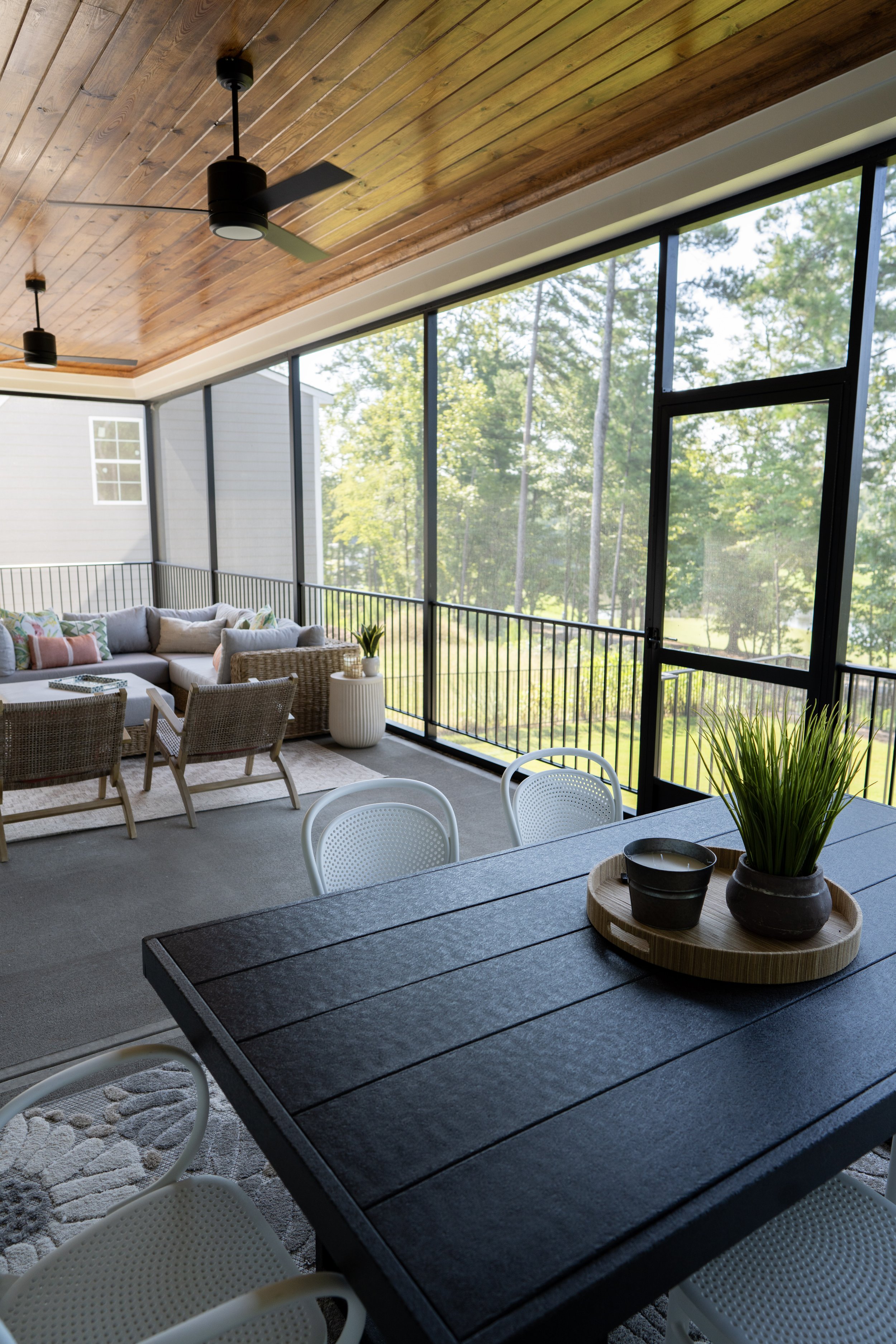 Enclosed porch with wood-paneled ceiling, ceiling fans, and large screened windows showing trees outside. Contains a black dining table with white chairs, a sofa with cushions, wicker chairs, and decorative plants.