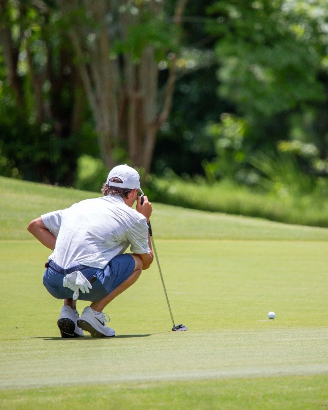 A man in a white shirt, blue shorts, and a white cap squatting on the green golf course, preparing to putt a golf ball with a putter.