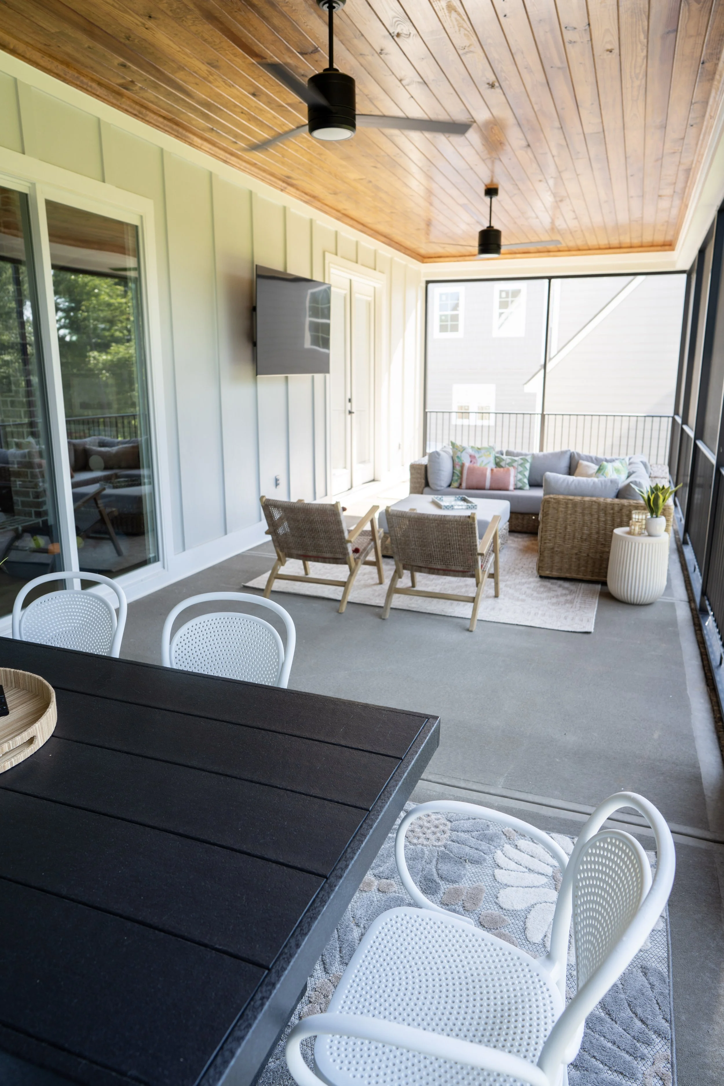 Covered balcony with black dining table and white chairs in the foreground, gray outdoor sofa with colorful pillows, two woven chairs, a television on the wall, wooden ceiling with two pendant lights, sliding glass door, and a railing with a view of 