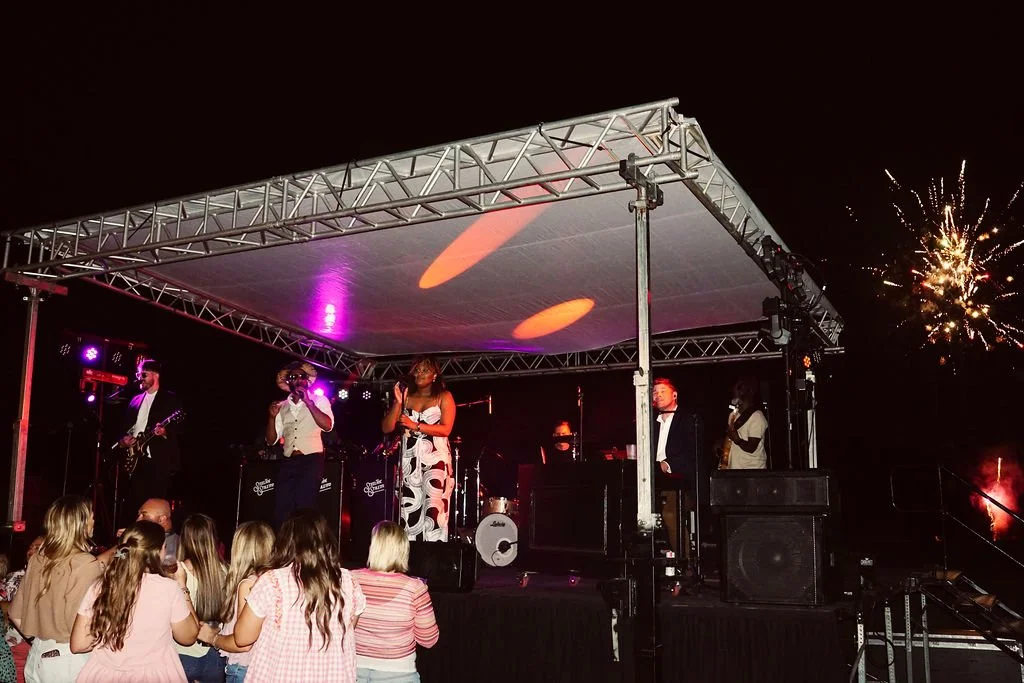 A live outdoor concert at night with a band performing on stage under a canopy, fireworks in the sky, and an audience of children watching.