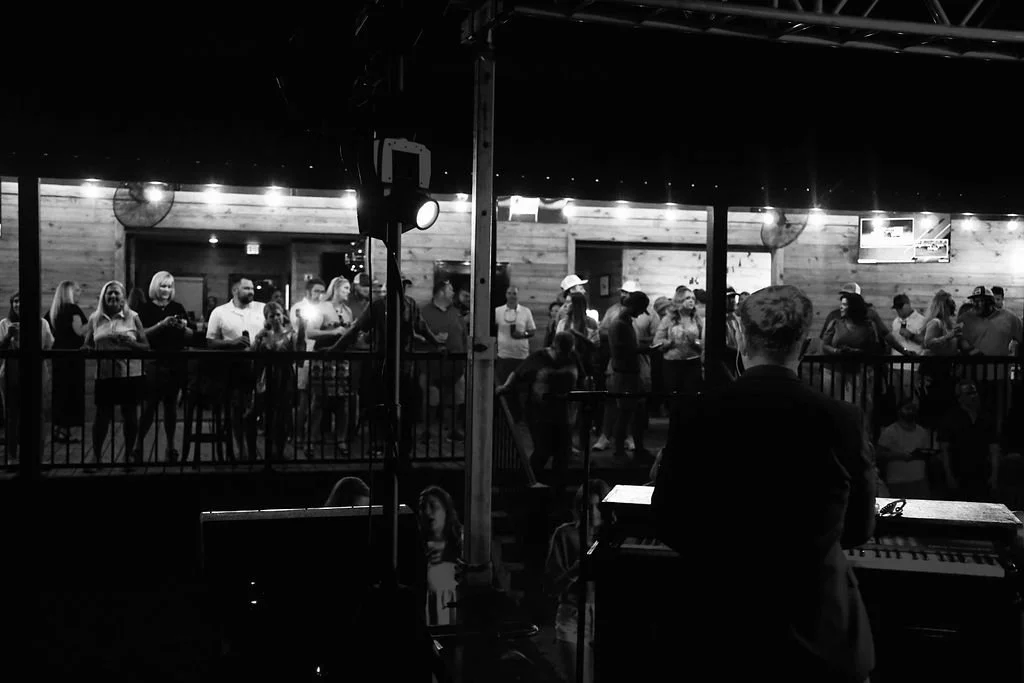 People gathered at an indoor concert or event, seen from behind a musician on stage. The audience is standing along a railing, with a rustic wooden wall and ceiling with string lights in the background.