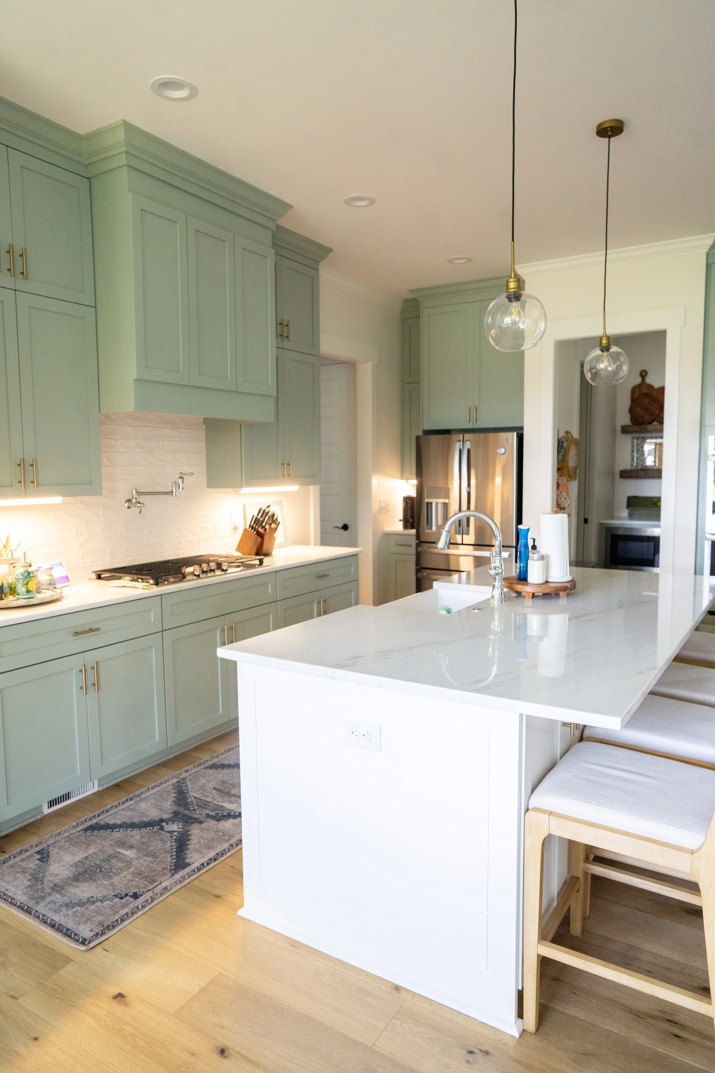 Modern kitchen with mint green cabinets, white marble island, stainless steel refrigerator, and pendant lighting.