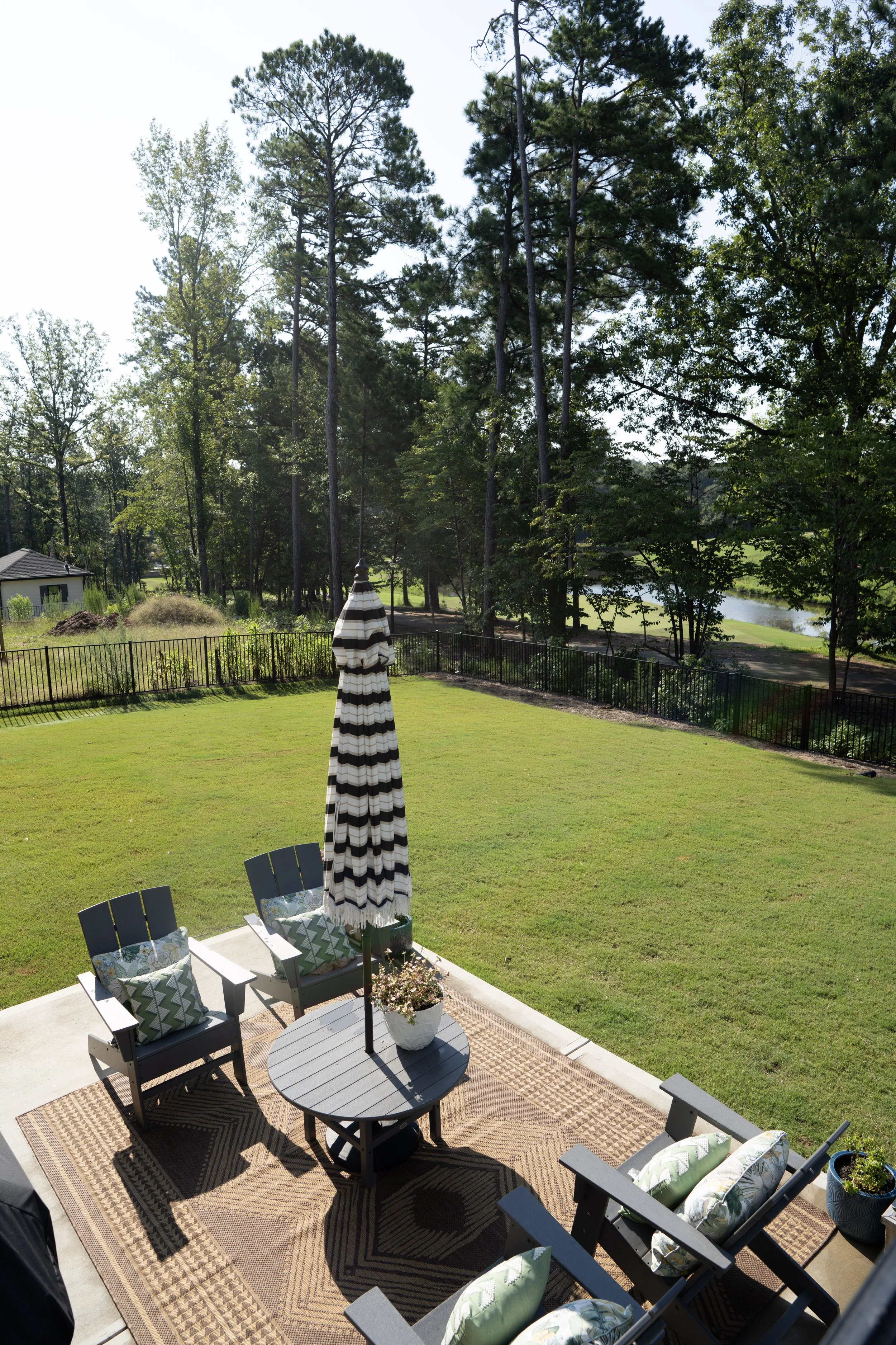 Backyard patio with a round table, chairs with pillows, a striped patio umbrella, and outdoor decor, overlooking a grassy yard, a fence, trees, and a river or lake in the background.