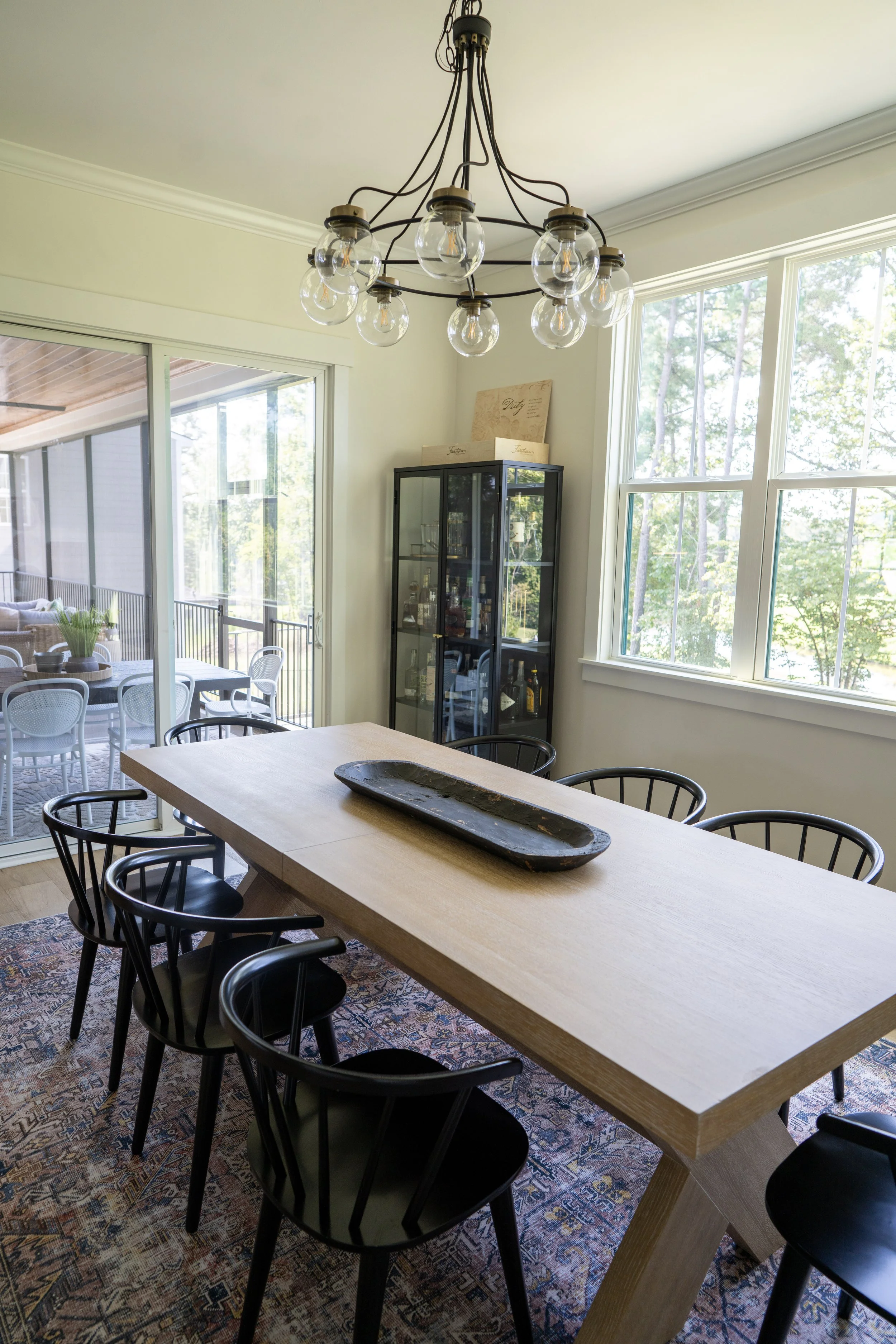 A dining room with a wooden table, six black chairs, a chandelier with exposed bulbs, a glass cabinet with bottles, large windows, and an outdoor patio visible through a sliding glass door.