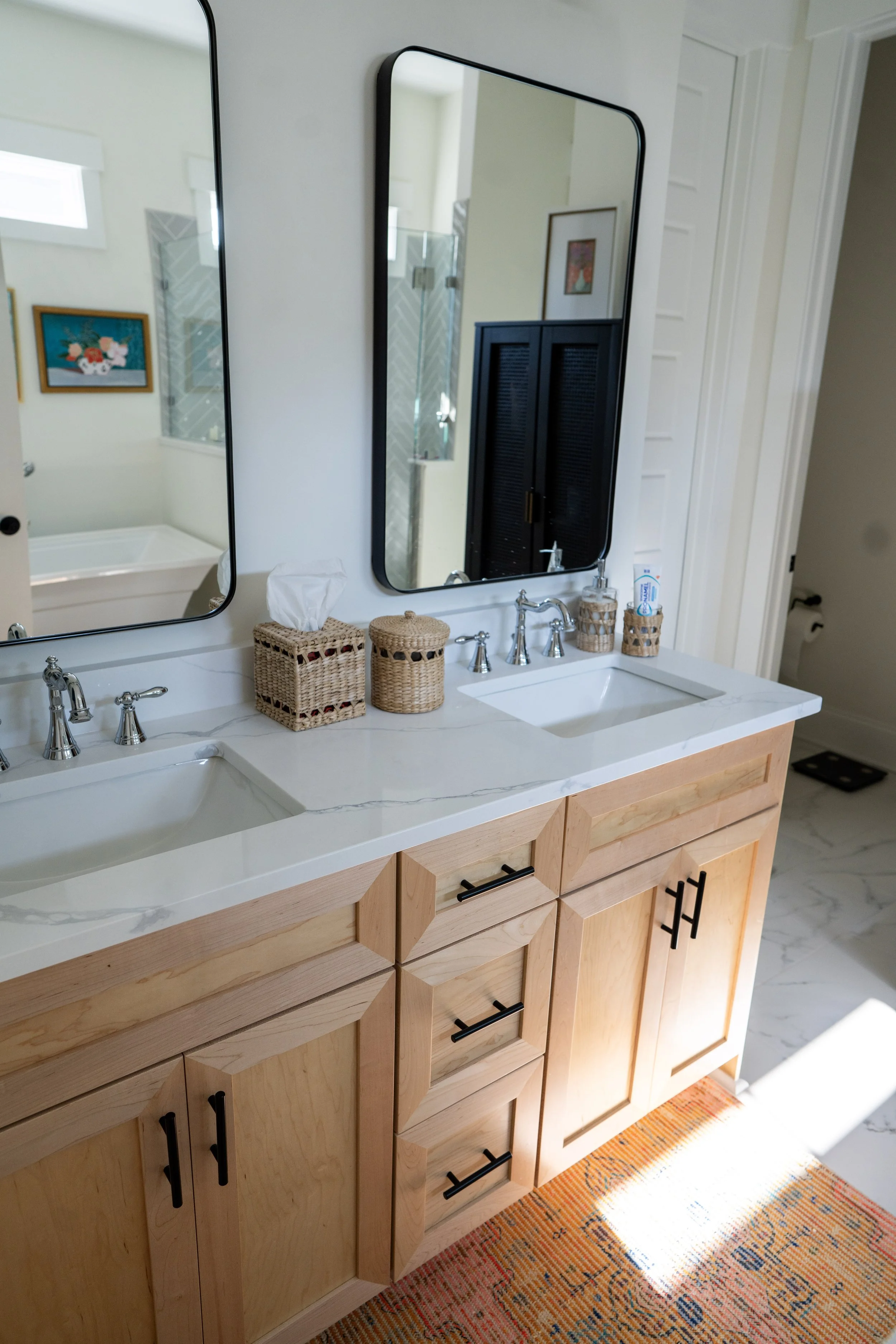 Bathroom vanity with two mirrors, wooden cabinets, marble countertop, two sinks with chrome fixtures, tissue box, jars, and toothbrush holder, with a colorful rug on the floor and partial view of a shower and wall art reflected in the mirrors.