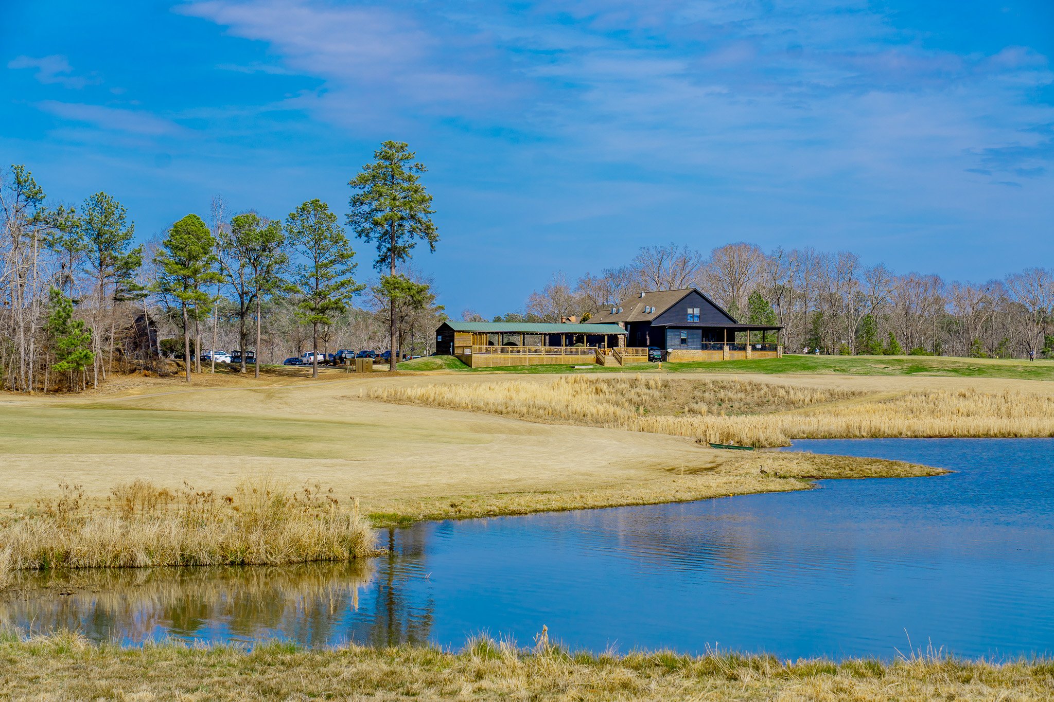 A golf course with a pond in the foreground, a house on a hill in the background, trees, and a blue sky.