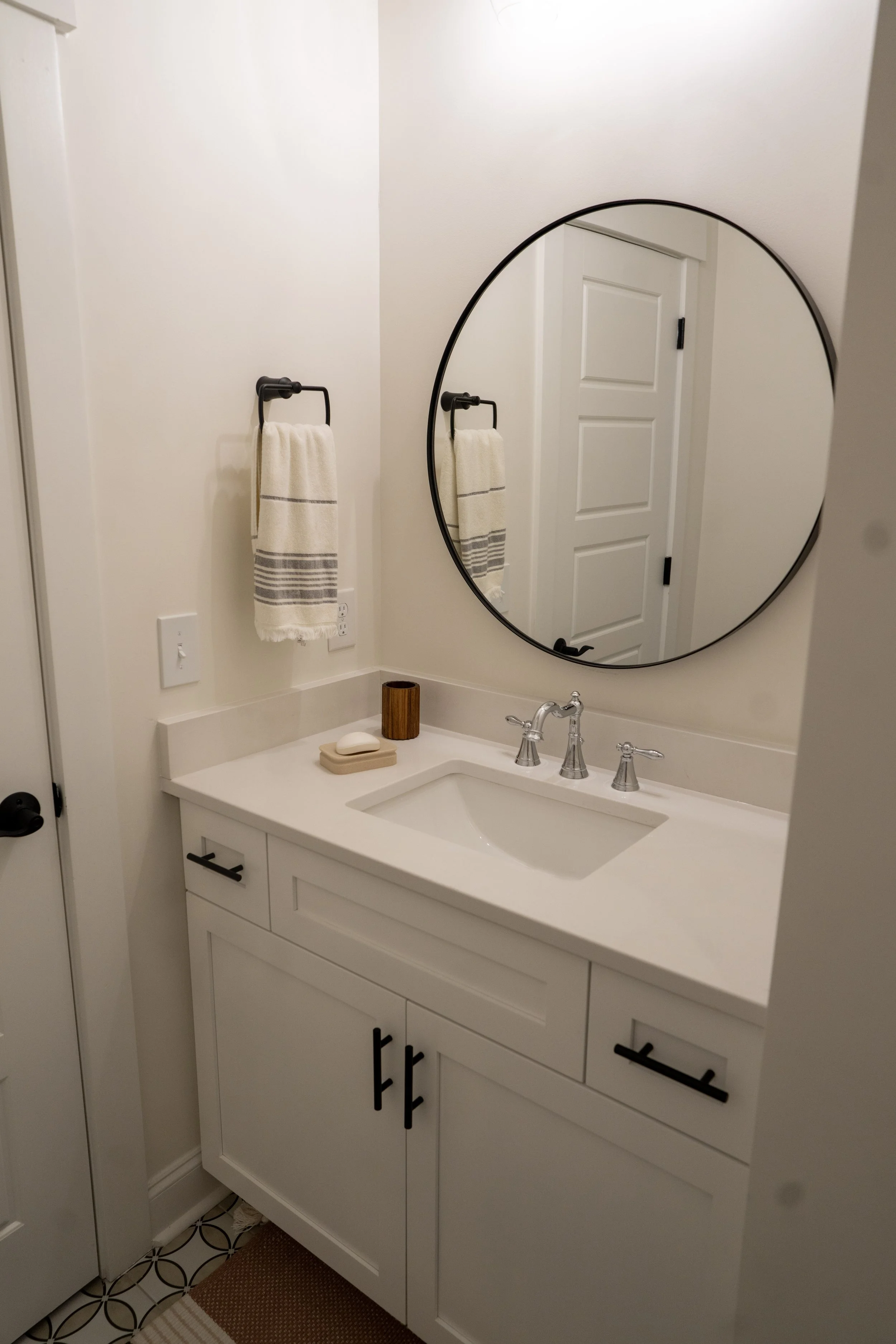 A bathroom vanity with a white cabinet, a rectangular sink, and a silver faucet, with a round mirror above it, a towel hanging on the wall, and toiletries on the countertop.