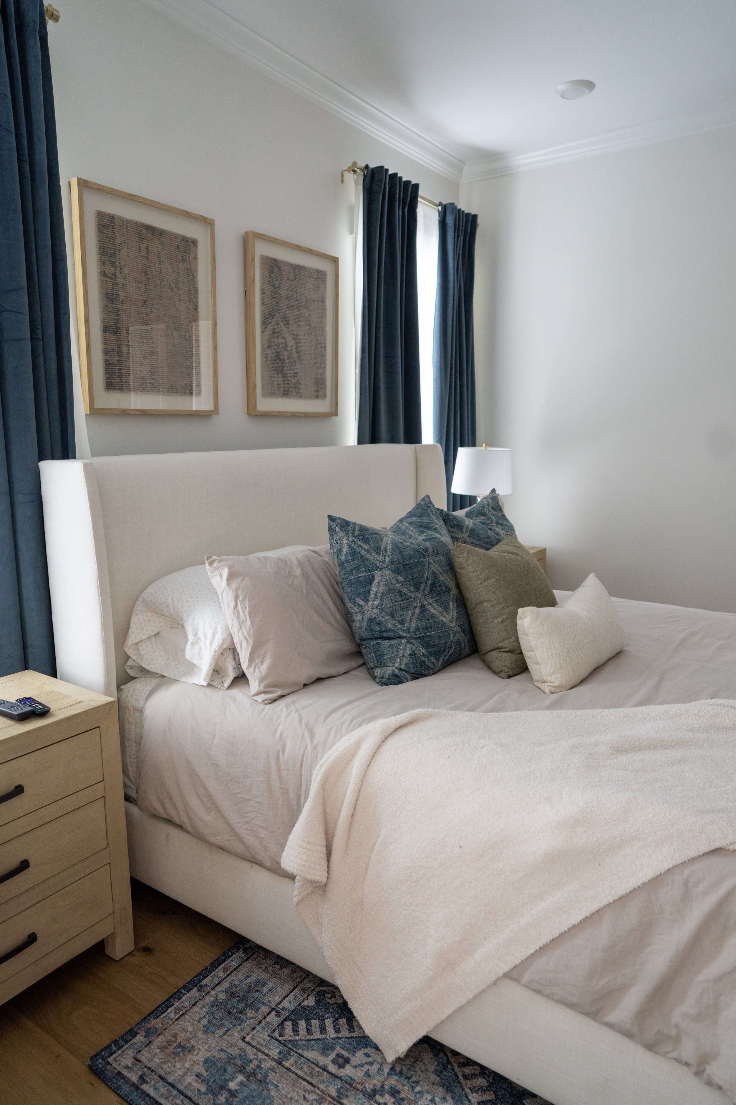 A neatly made bed with beige and blue pillows in a bedroom with white walls, navy blue curtains, a nightstand with a remote, and framed artwork overhead.
