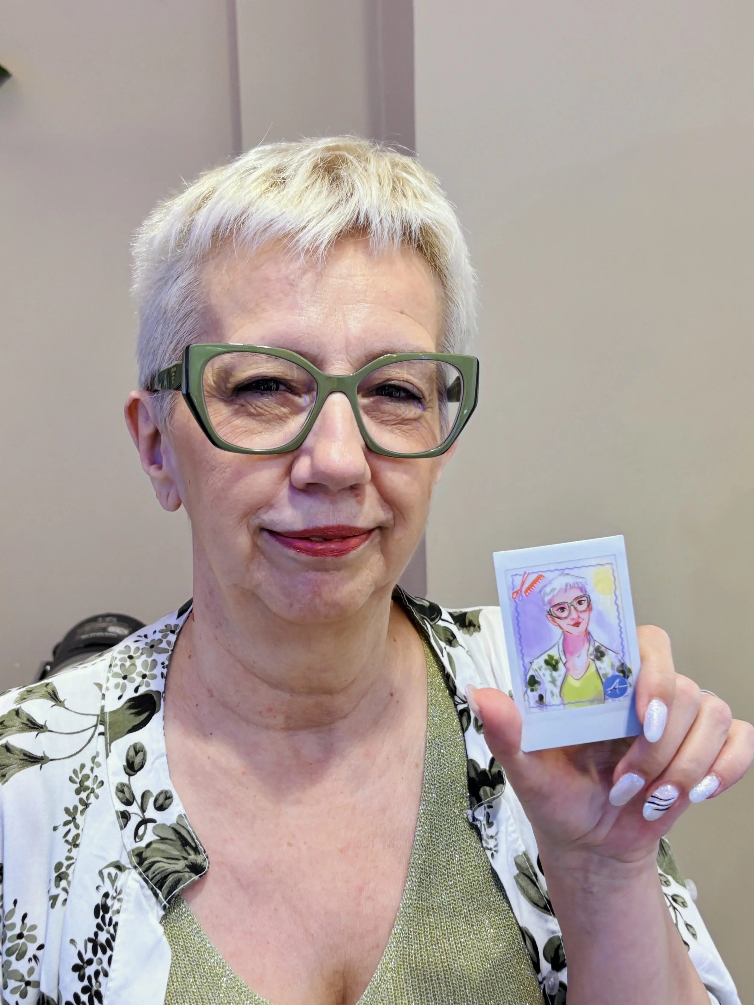 A woman with short blonde hair and large green glasses smiling while holding a colorful illustrated portrait of herself.