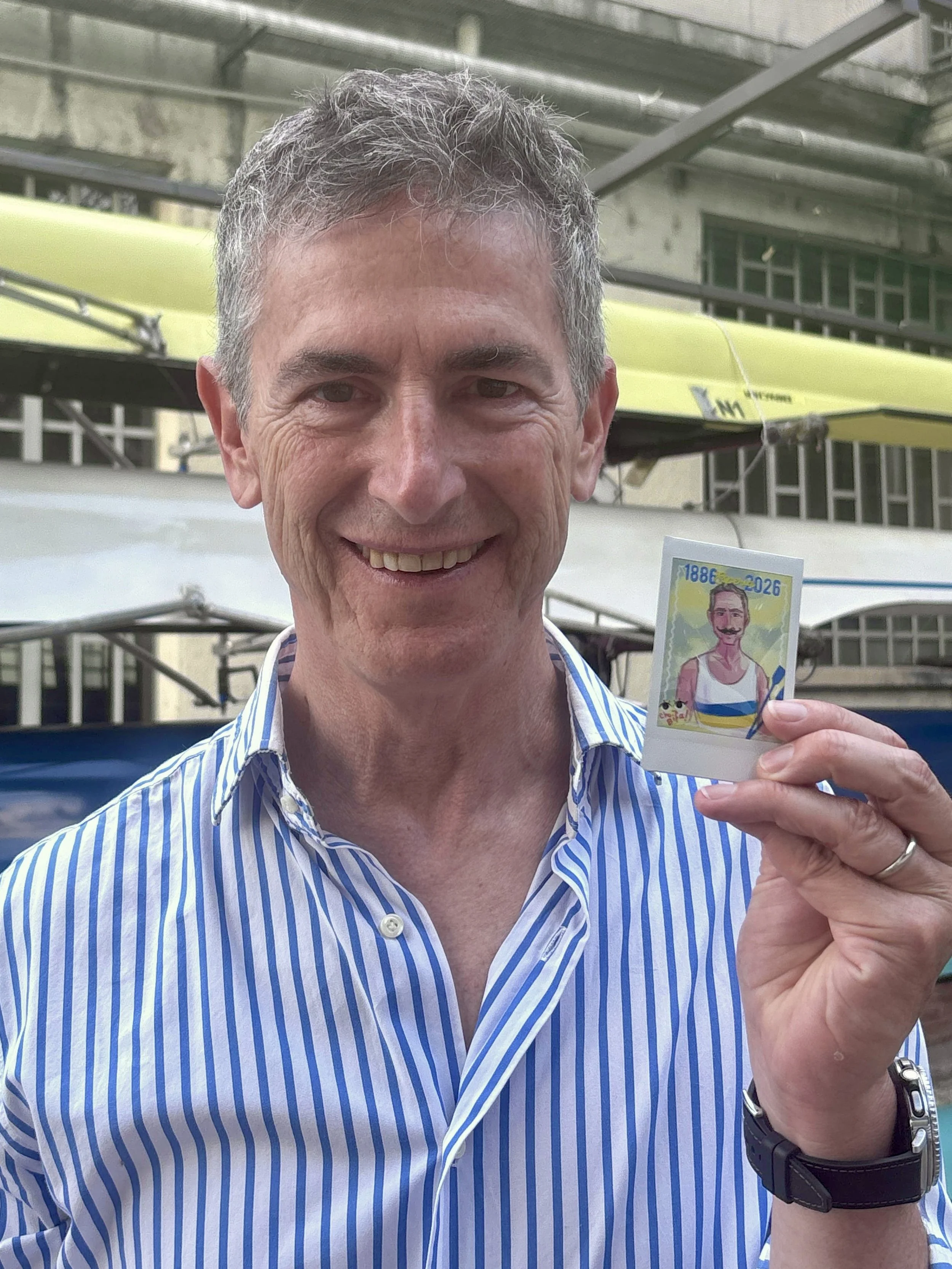 A smiling middle-aged man with gray curly hair, wearing a blue and white striped shirt, holding a colorful cartoon-style postcard with a smiling cartoon of himself. The background shows a yellow boat and some metal scaffolding.