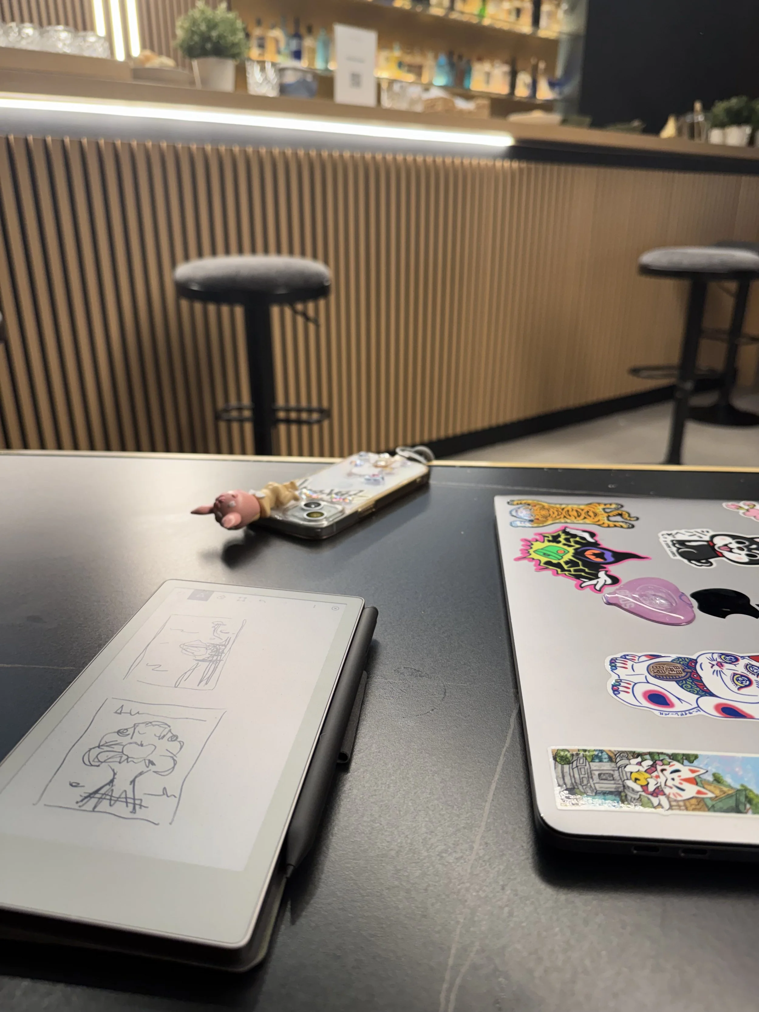A black table with a white tablet displaying a sketch, a closed laptop with stickers, and a phone with a pig keychain. The background features a bar counter with bar stools, a wooden slat front, and some plants and bottles on shelves.